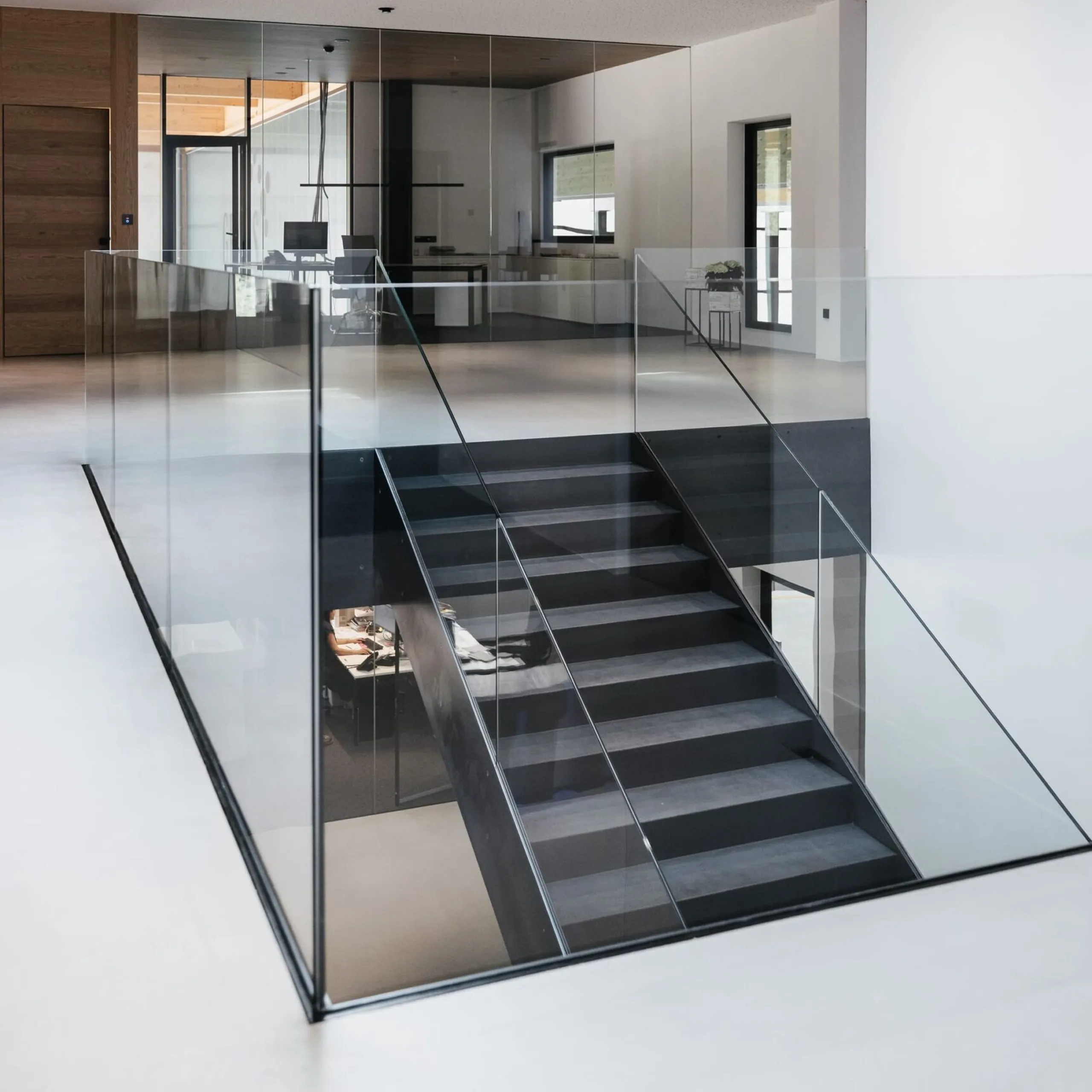 Heller moderner Bürobereich mit Glasgeländer, schwarzer Metalltreppe, weißem Boden und Oberlichtfenster; Glaswände und Arbeitsplätze im Hintergrund.