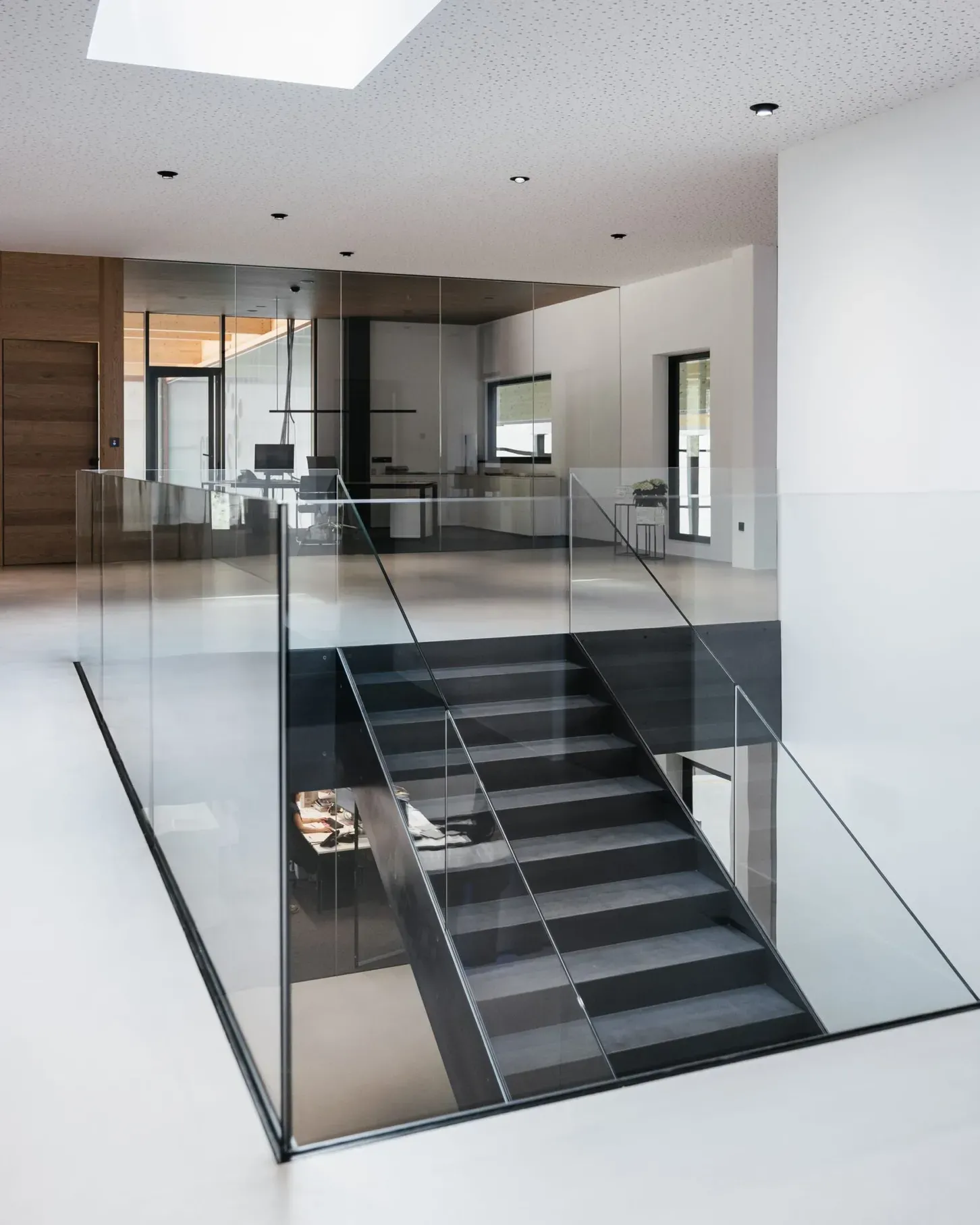 Heller moderner Bürobereich mit Glasgeländer, schwarzer Metalltreppe, weißem Boden und Oberlichtfenster; Glaswände und Arbeitsplätze im Hintergrund.