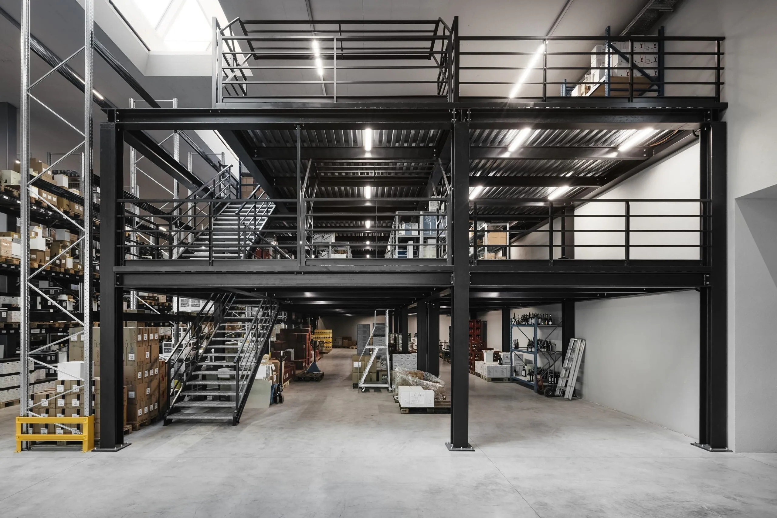 Große Lagerhalle mit schwarzer Stahlkonstruktion, Zwischenebene, Treppen, Geländer, Regale und Paletten – Stahlbau in Südtirol.