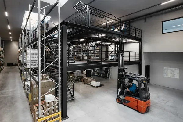 Große Lagerhalle eines Südtiroler Stahlbaubetriebs: schwarze Metallkonstruktion mit Mezzanin, Regale voller Kartons, Gabelstapler mit Fahrer im Vordergrund.