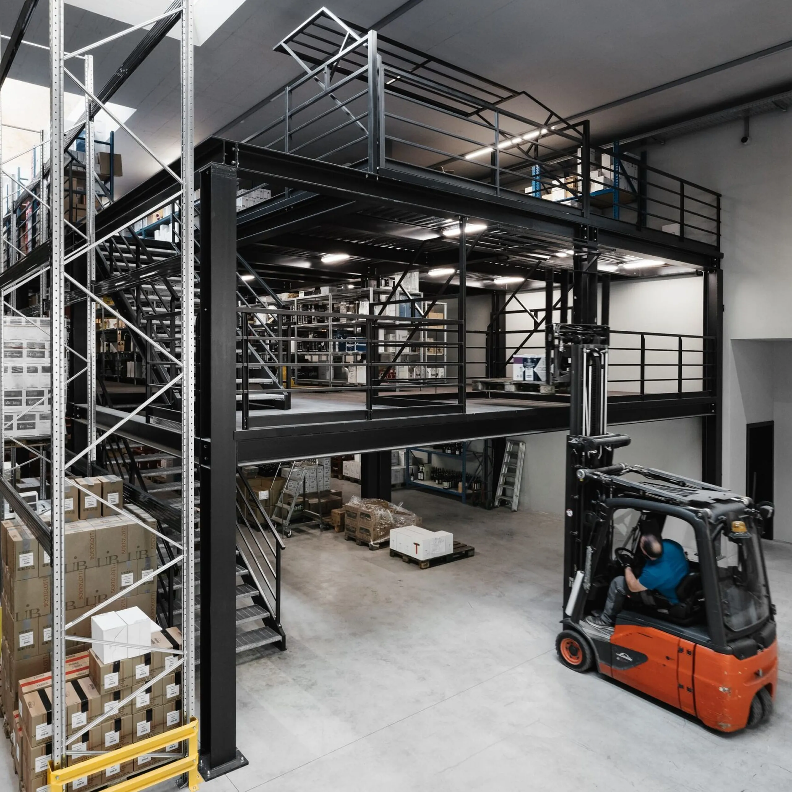 Große Lagerhalle eines Südtiroler Stahlbaubetriebs: schwarze Metallkonstruktion mit Mezzanin, Regale voller Kartons, Gabelstapler mit Fahrer im Vordergrund.