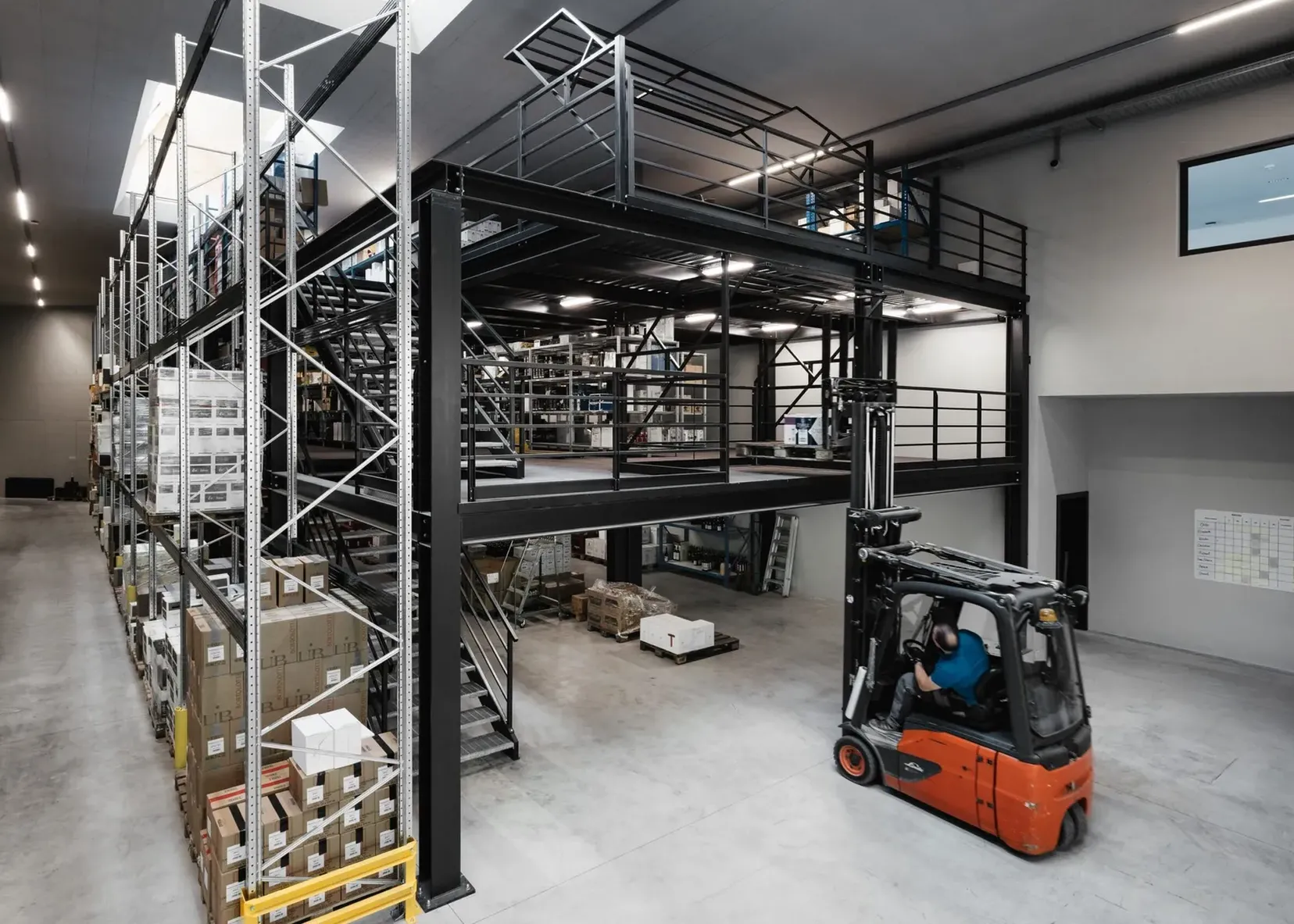 Große Lagerhalle eines Südtiroler Stahlbaubetriebs: schwarze Metallkonstruktion mit Mezzanin, Regale voller Kartons, Gabelstapler mit Fahrer im Vordergrund.