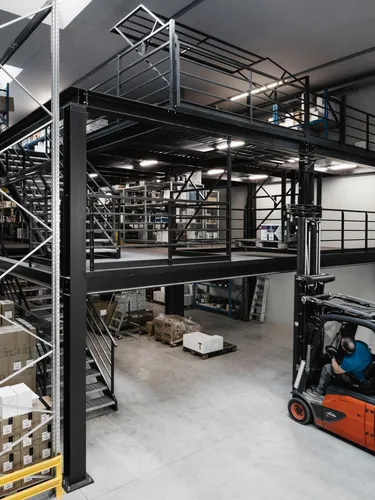Große Lagerhalle eines Südtiroler Stahlbaubetriebs: schwarze Metallkonstruktion mit Mezzanin, Regale voller Kartons, Gabelstapler mit Fahrer im Vordergrund.