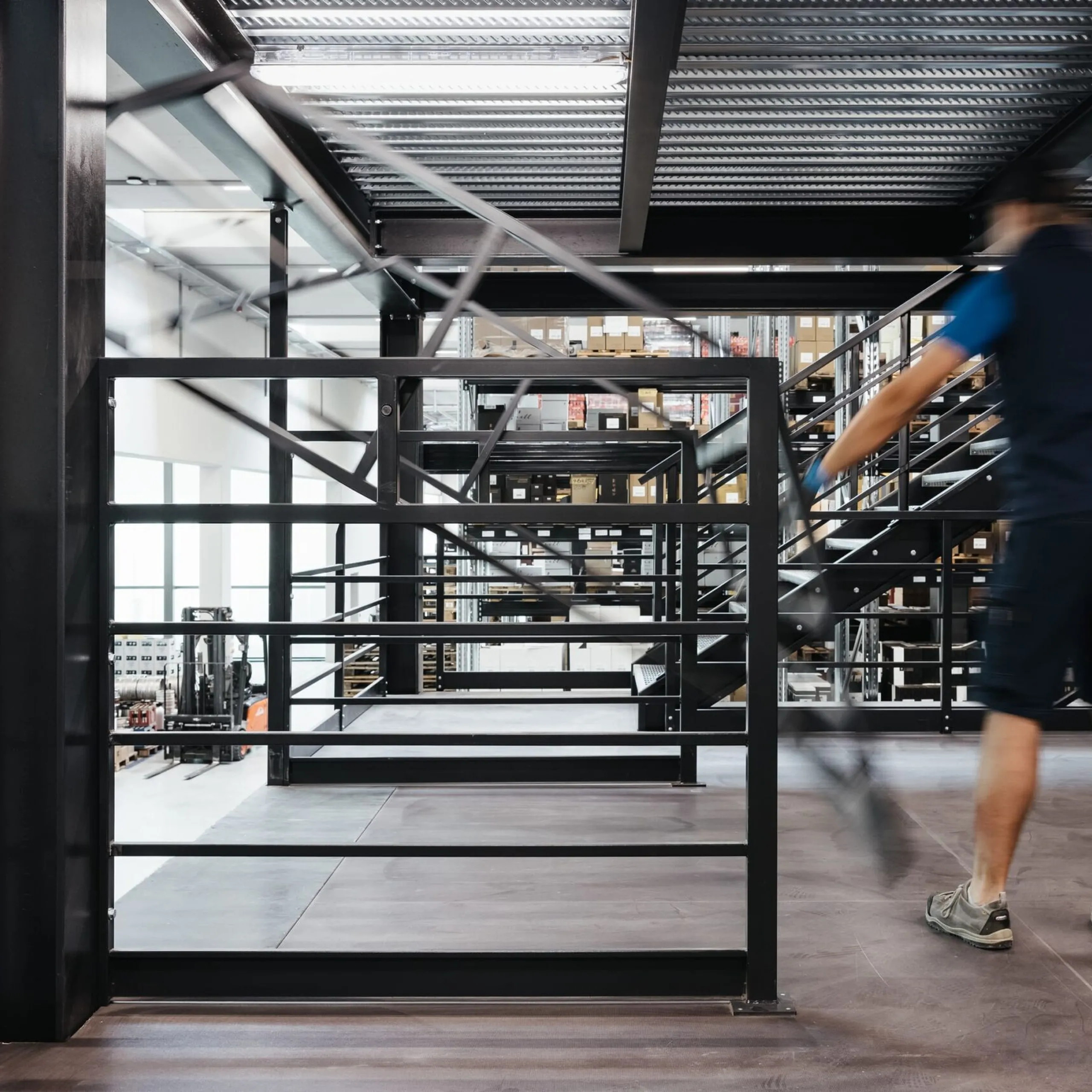 Industriehalle eines Metallbau-Unternehmens in Südtirol; eine Person in Arbeitskleidung geht über eine Stahlgalerie, Geländer und Lager im Hintergrund.