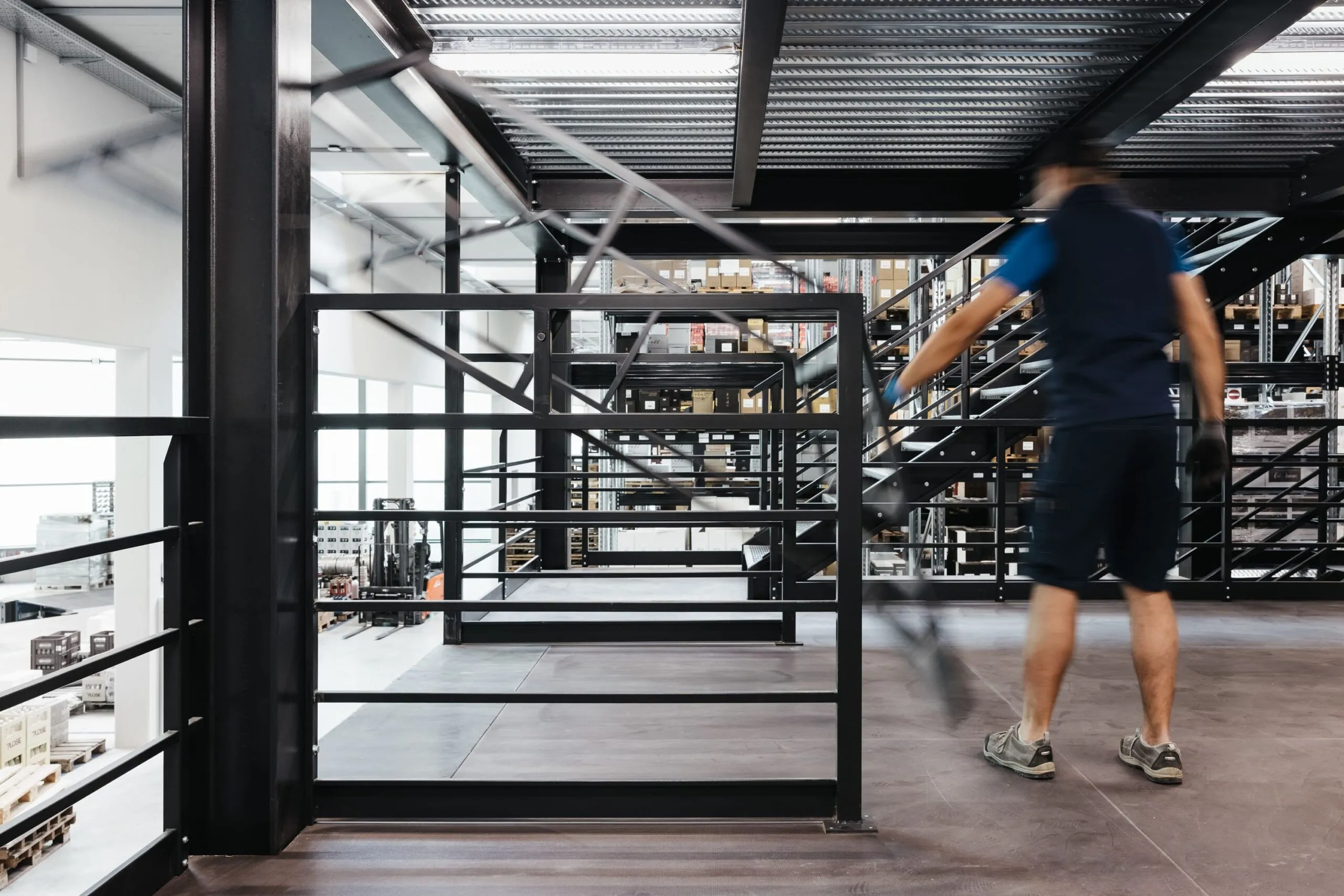 Industriehalle eines Metallbau-Unternehmens in Südtirol; eine Person in Arbeitskleidung geht über eine Stahlgalerie, Geländer und Lager im Hintergrund.