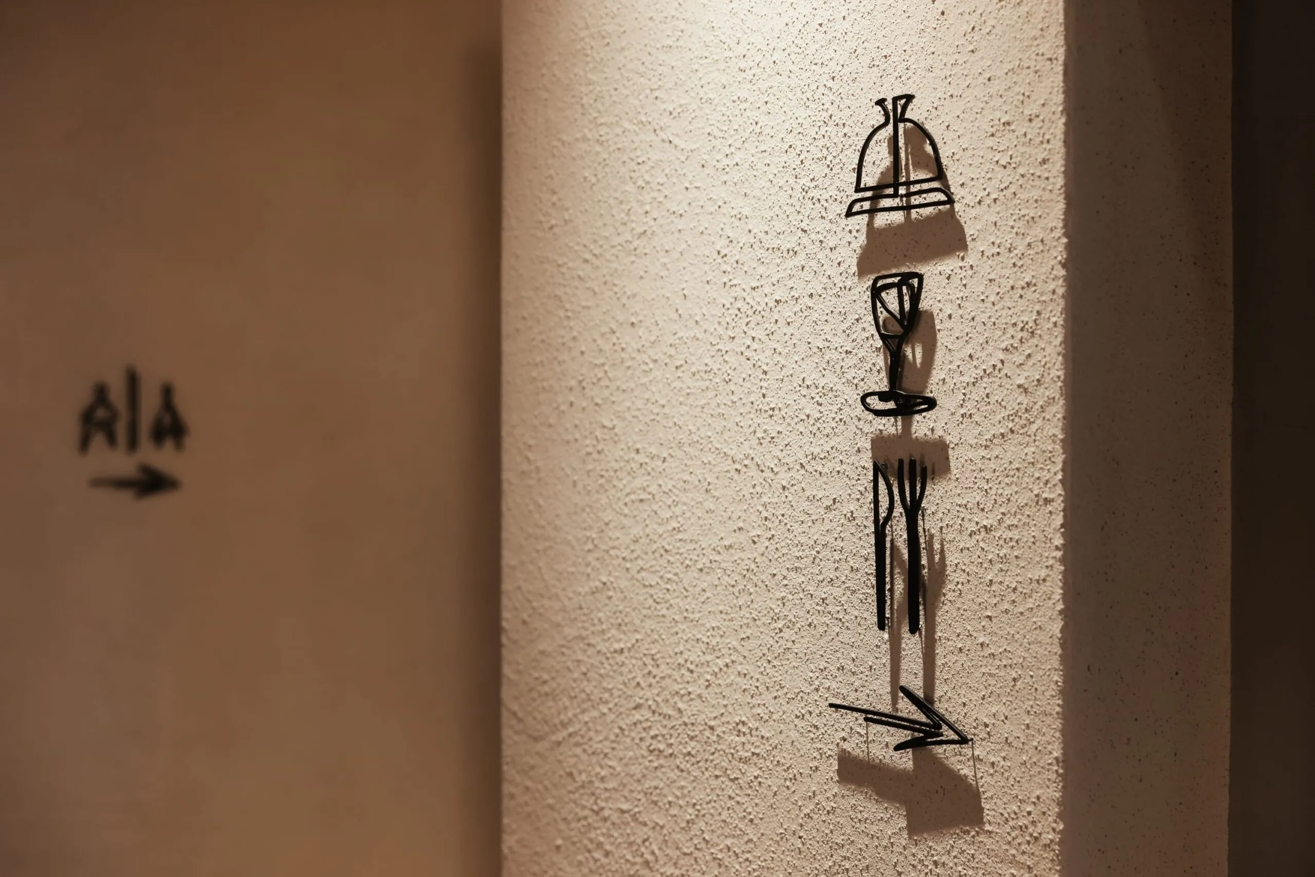 Colonna di muro ruvido con graffiti neri stilizzati allineati verticalmente; sullo sfondo sinistro, simbolo AIA sfocato con freccia.