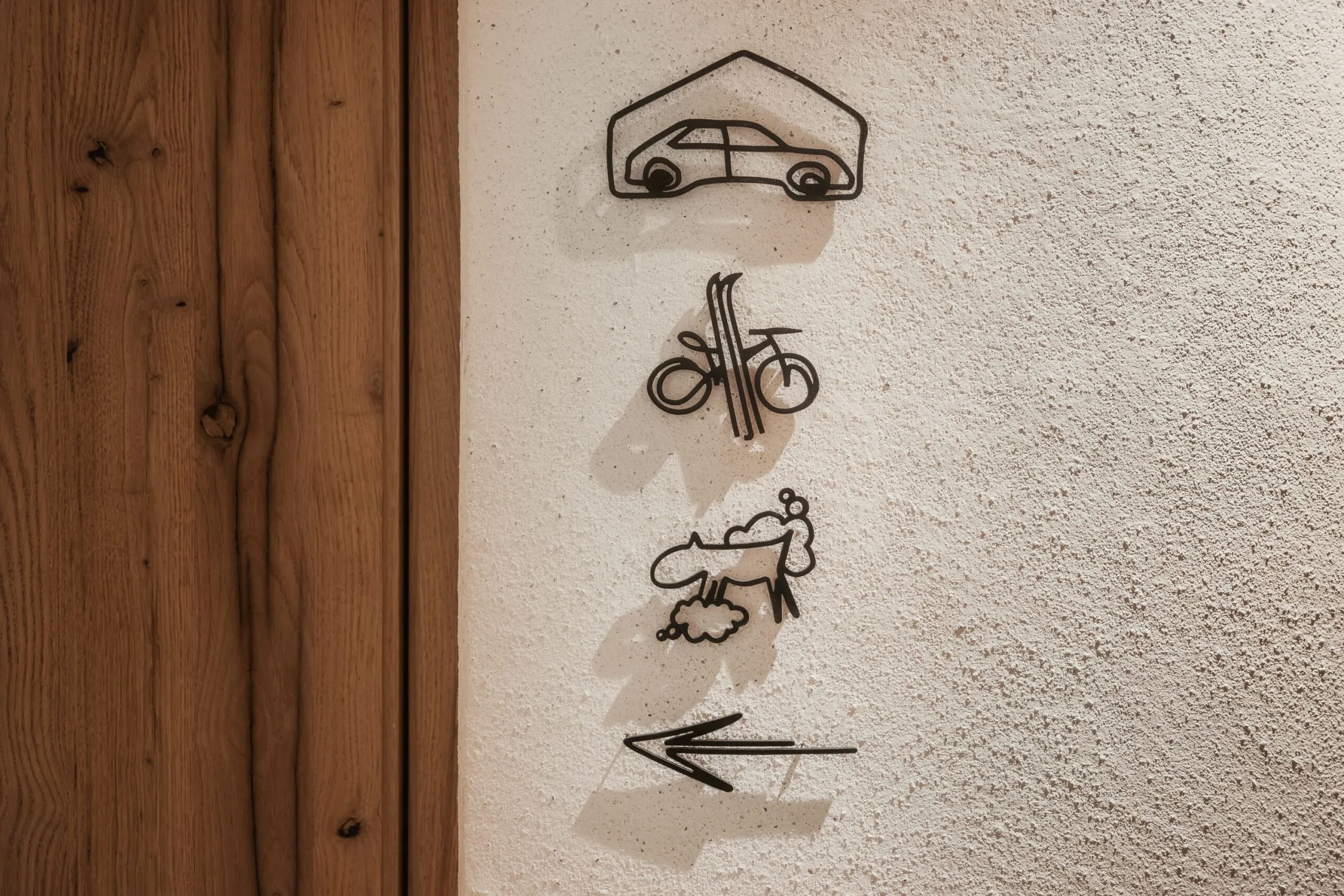 Serie verticale di icone nere su parete chiara accanto a una porta in legno: auto sotto tetto, bici stilizzata, figura astratta con vapore, freccia verso sinistra.