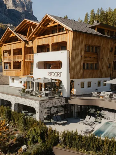 Hotel in legno stile chalet con piscina e terrazza all'aperto, situato su pendio montano con foreste autunnali e montagne sullo sfondo.