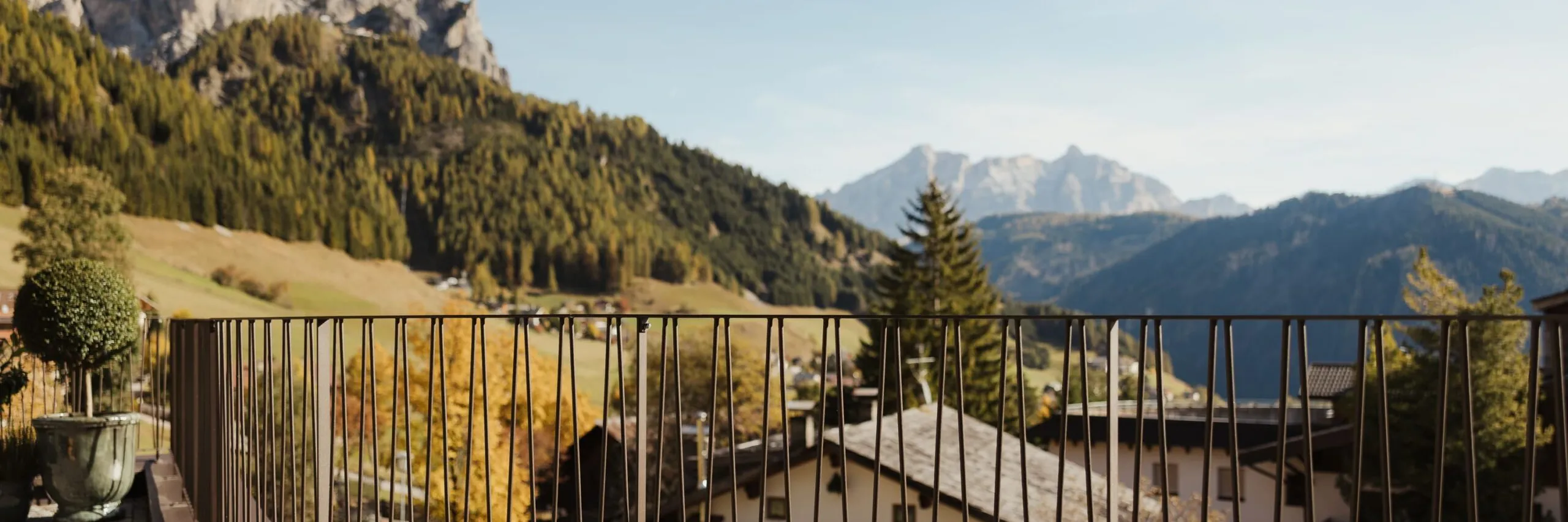 Balconcino con corrimano metallico curvo, vista su paesaggio alpino sudtirolese: case, piscina e alberi autunnali tra montagne lontane.