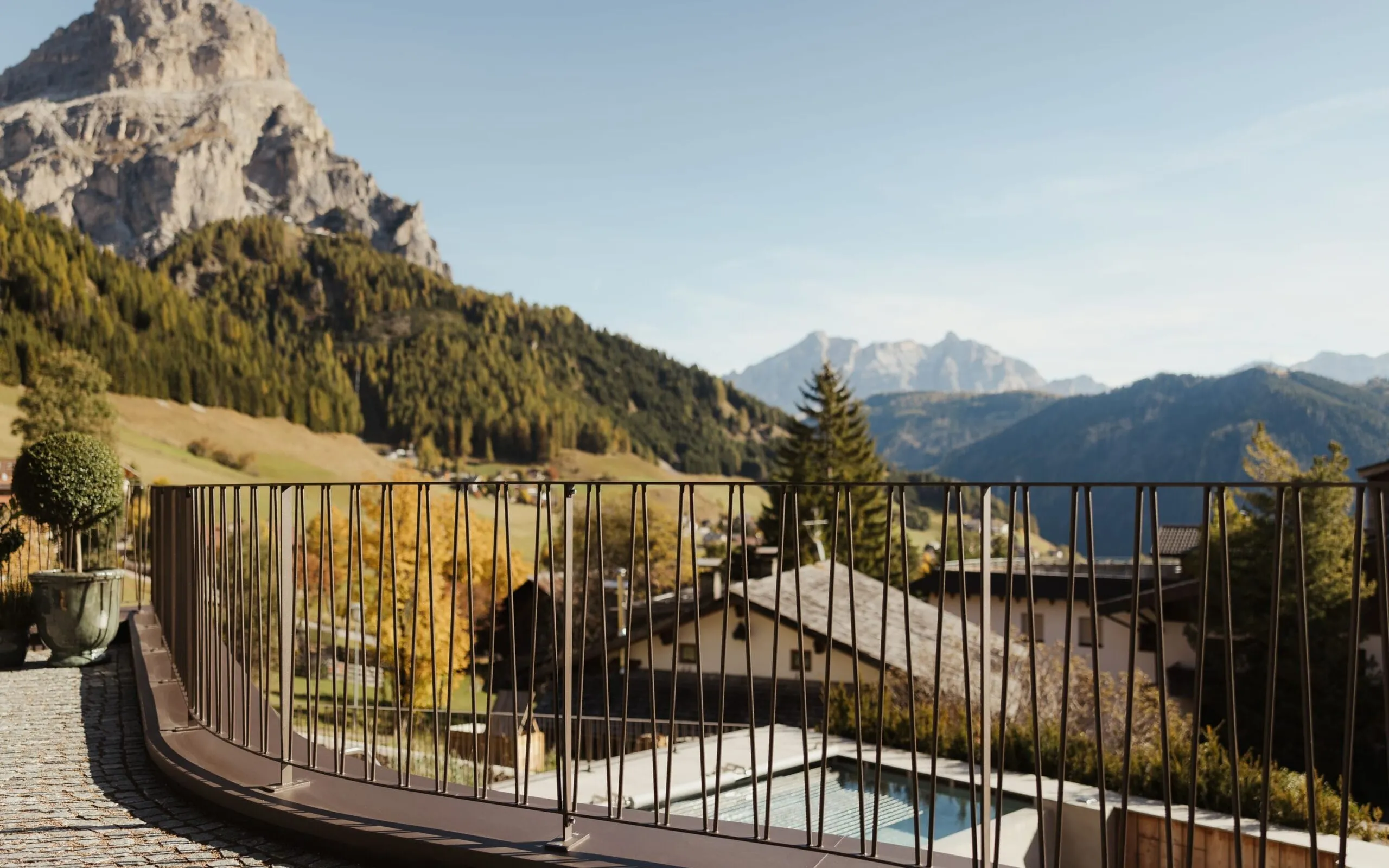 Balconcino con corrimano metallico curvo, vista su paesaggio alpino sudtirolese: case, piscina e alberi autunnali tra montagne lontane.
