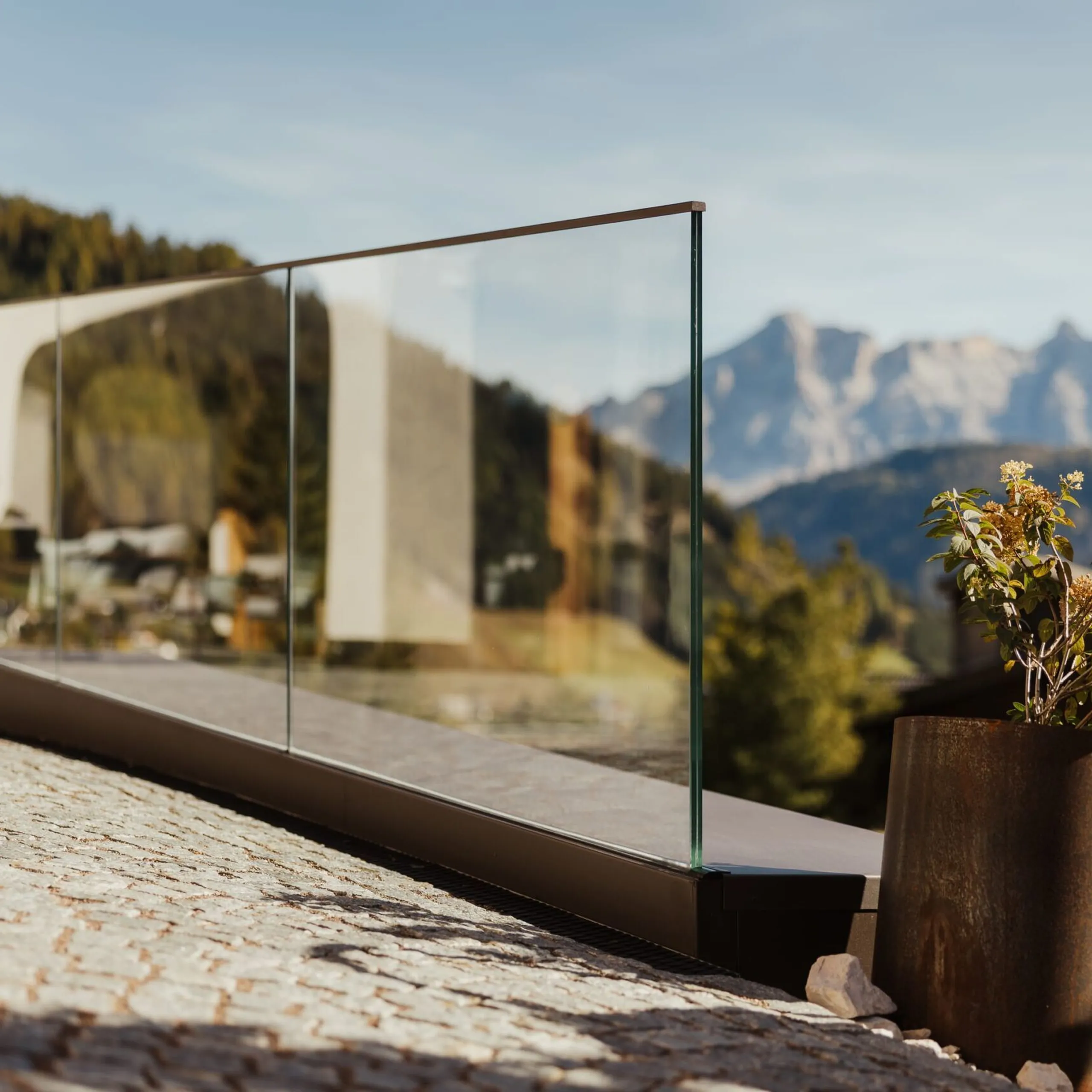 Balustrata in vetro su terrazzo in pietra con vaso metallico arrugginito e fiori; sullo sfondo montagne alpine e case.