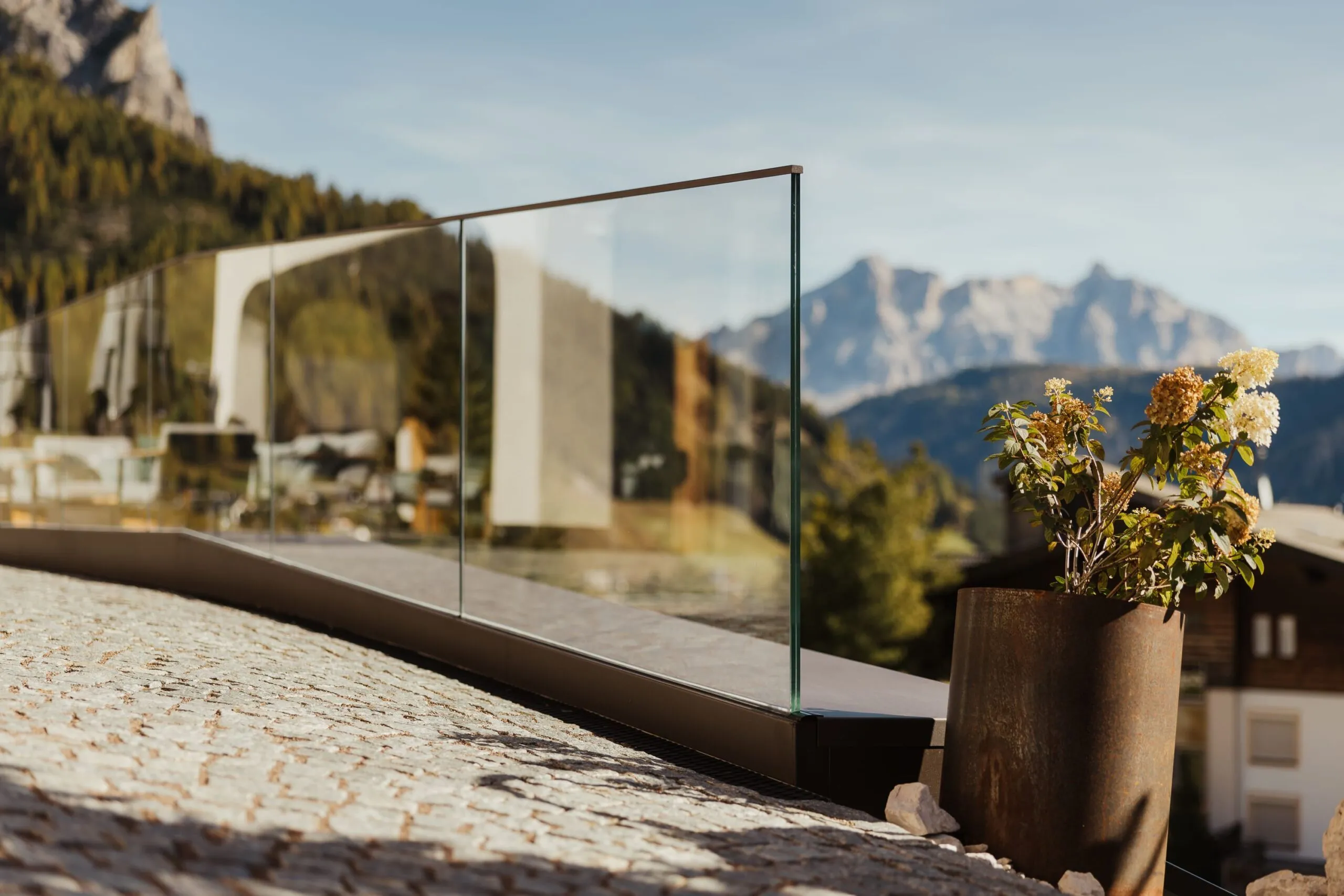 Balustrata in vetro su terrazzo in pietra con vaso metallico arrugginito e fiori; sullo sfondo montagne alpine e case.