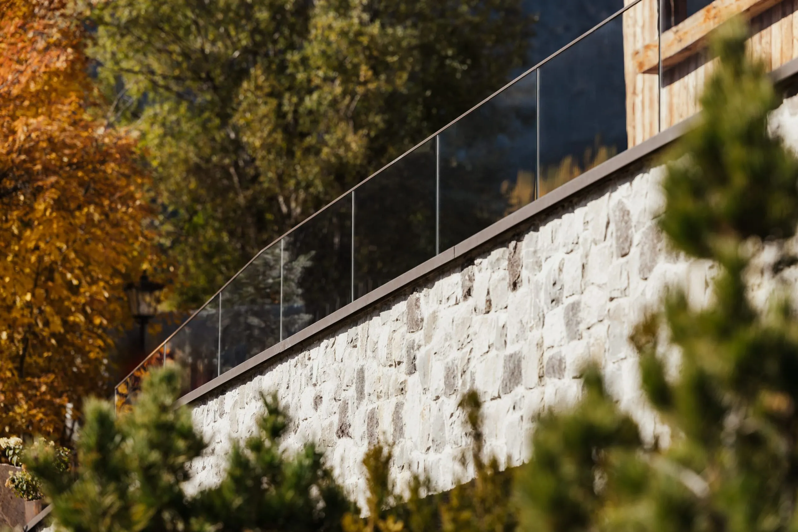 Parapetto in vetro con cornice metallica lungo un muro di pietra, sfondo di alberi autunnali e vegetazione in primo piano.
