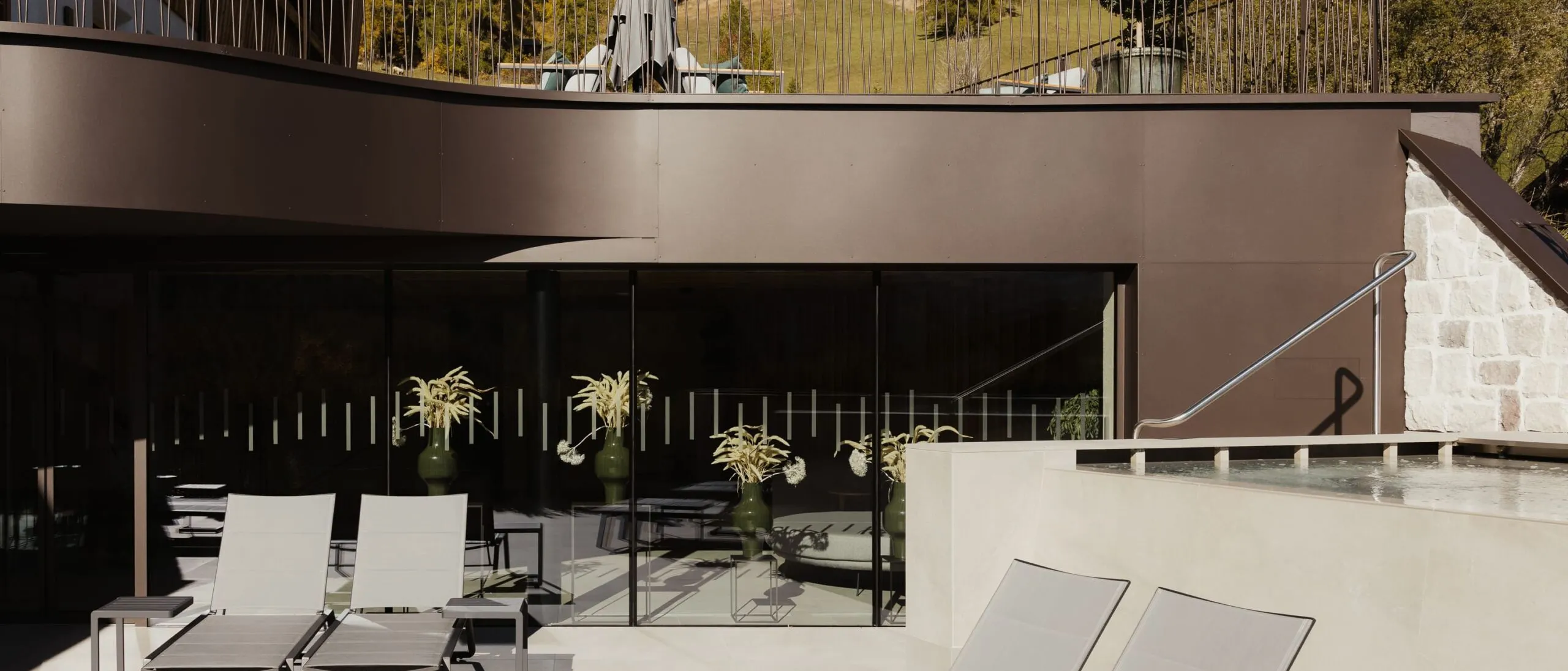 Patio esterno di un'azienda metallica in Alto Adige: lettini a sdraio grigie, tavolino, vetrata scorrevole, piante in vasi, balaustra e paesaggio boscoso.