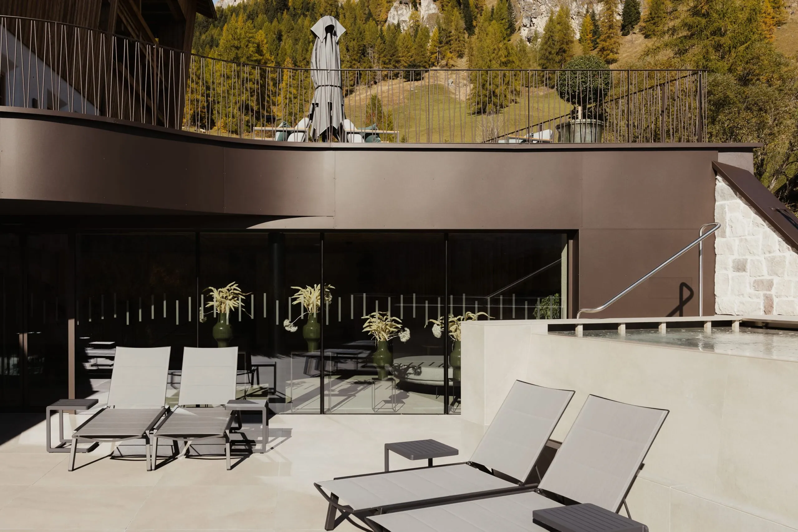 Patio esterno di un'azienda metallica in Alto Adige: lettini a sdraio grigie, tavolino, vetrata scorrevole, piante in vasi, balaustra e paesaggio boscoso.