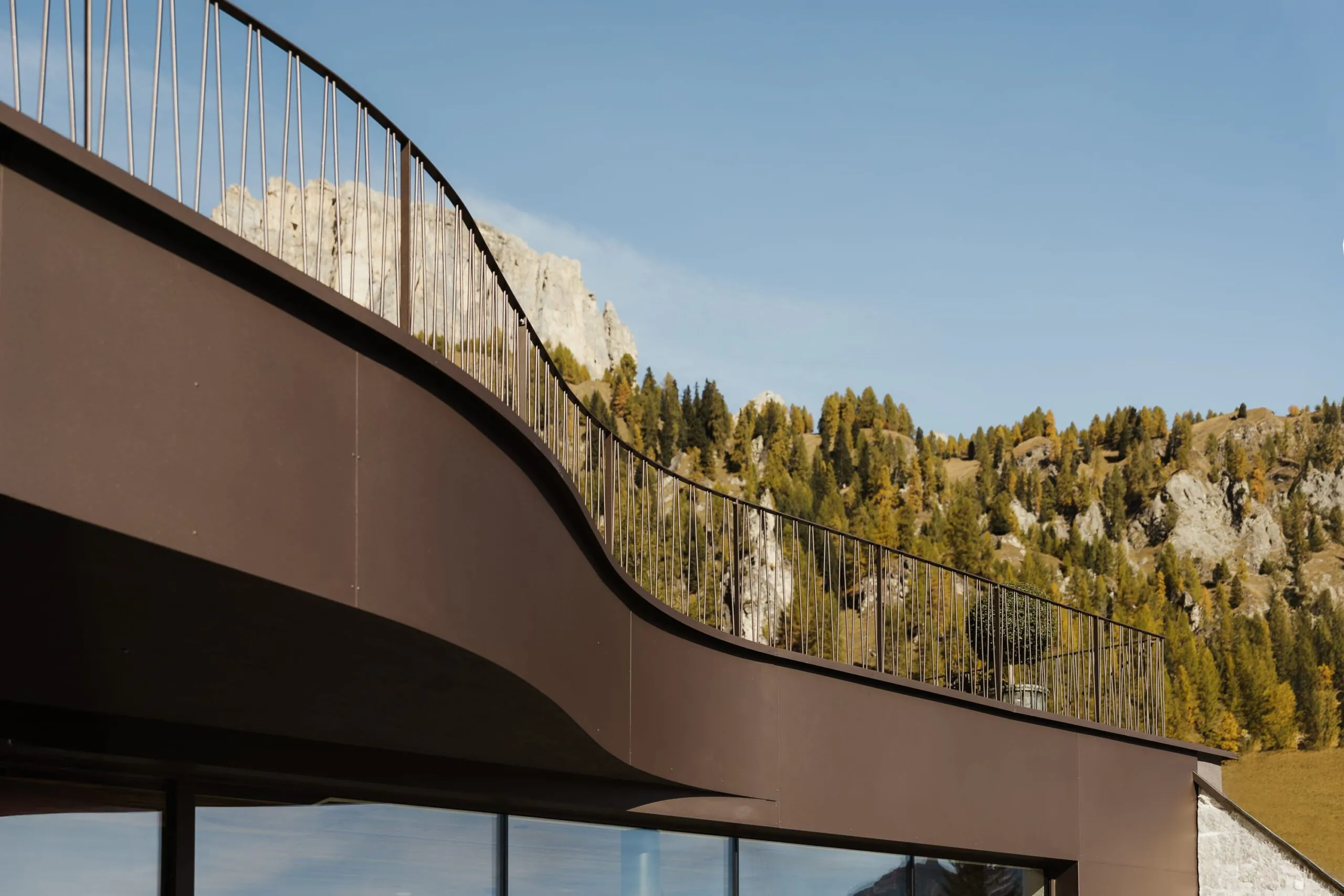 Balcone curvo in metallo marrone su edificio moderno, parapetti verticali; sfondo montuoso con alberi e rocce, cielo azzurro.