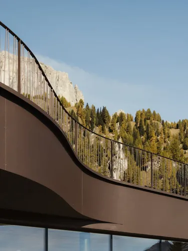 Balcone curvo in metallo marrone su edificio moderno, parapetti verticali; sfondo montuoso con alberi e rocce, cielo azzurro.