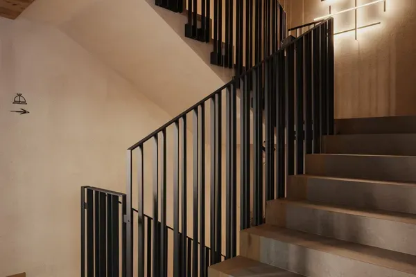 Scalinata interna con corrimanti in metallo neri, gradini in cemento, pareti beige e soffitto in legno; lampada moderna a croce sulla parete destra.