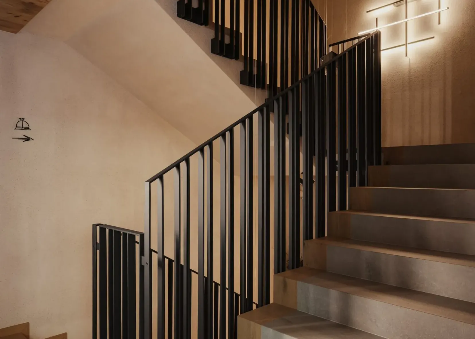 Scalinata interna con corrimanti in metallo neri, gradini in cemento, pareti beige e soffitto in legno; lampada moderna a croce sulla parete destra.