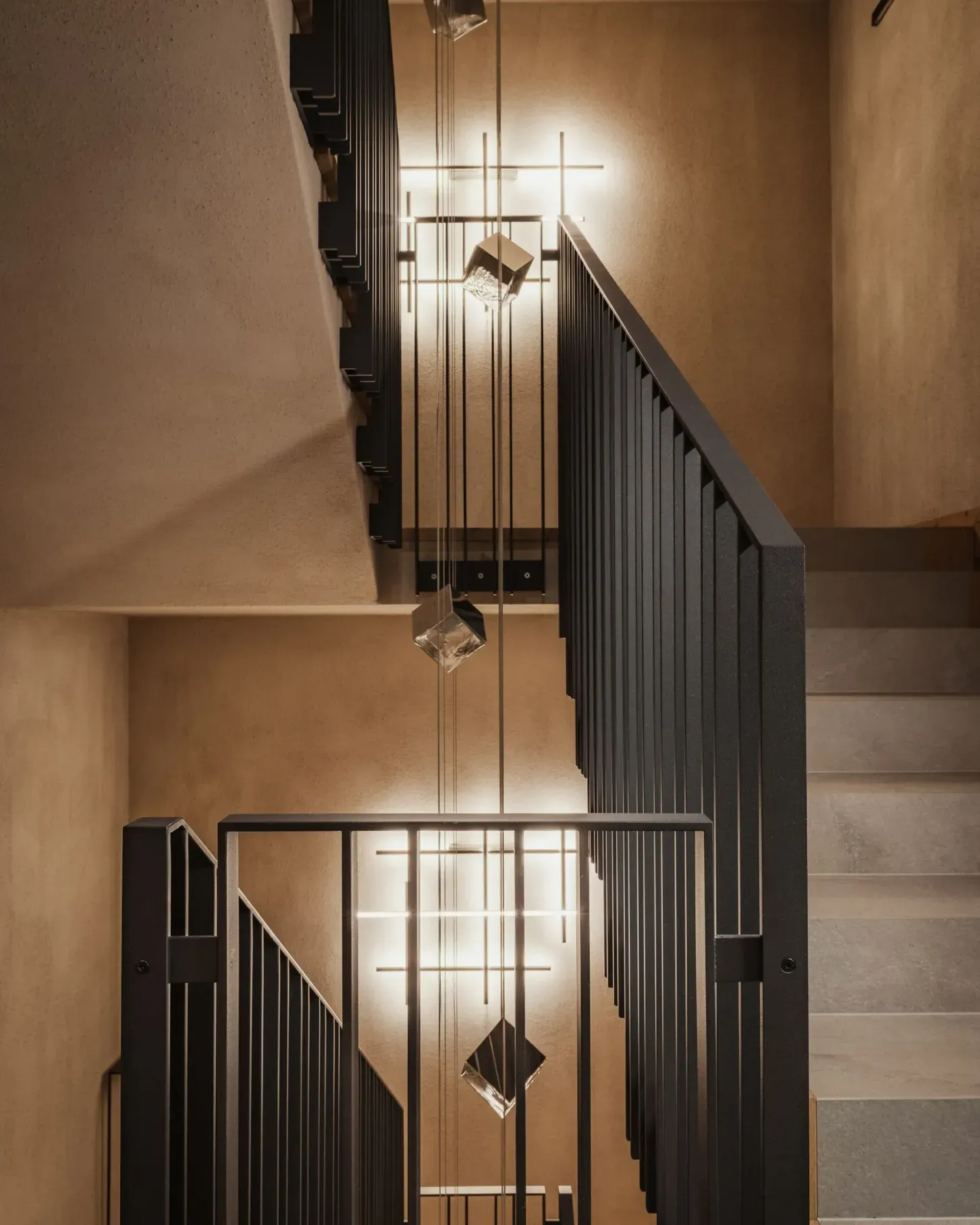 Scala interna in metallo nero con corrimano a bastoni e balaustre verticali, pareti beige, illuminata da lampade pendenti moderne con cubi di vetro.