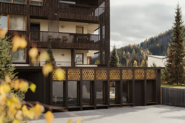 Mehrstöckiges Gebäude mit dunkler Holzfassade und Balkonen; untere Etage mit Gitterverkleidung, davor gelbe Herbstblätter und Wald im Hintergrund.