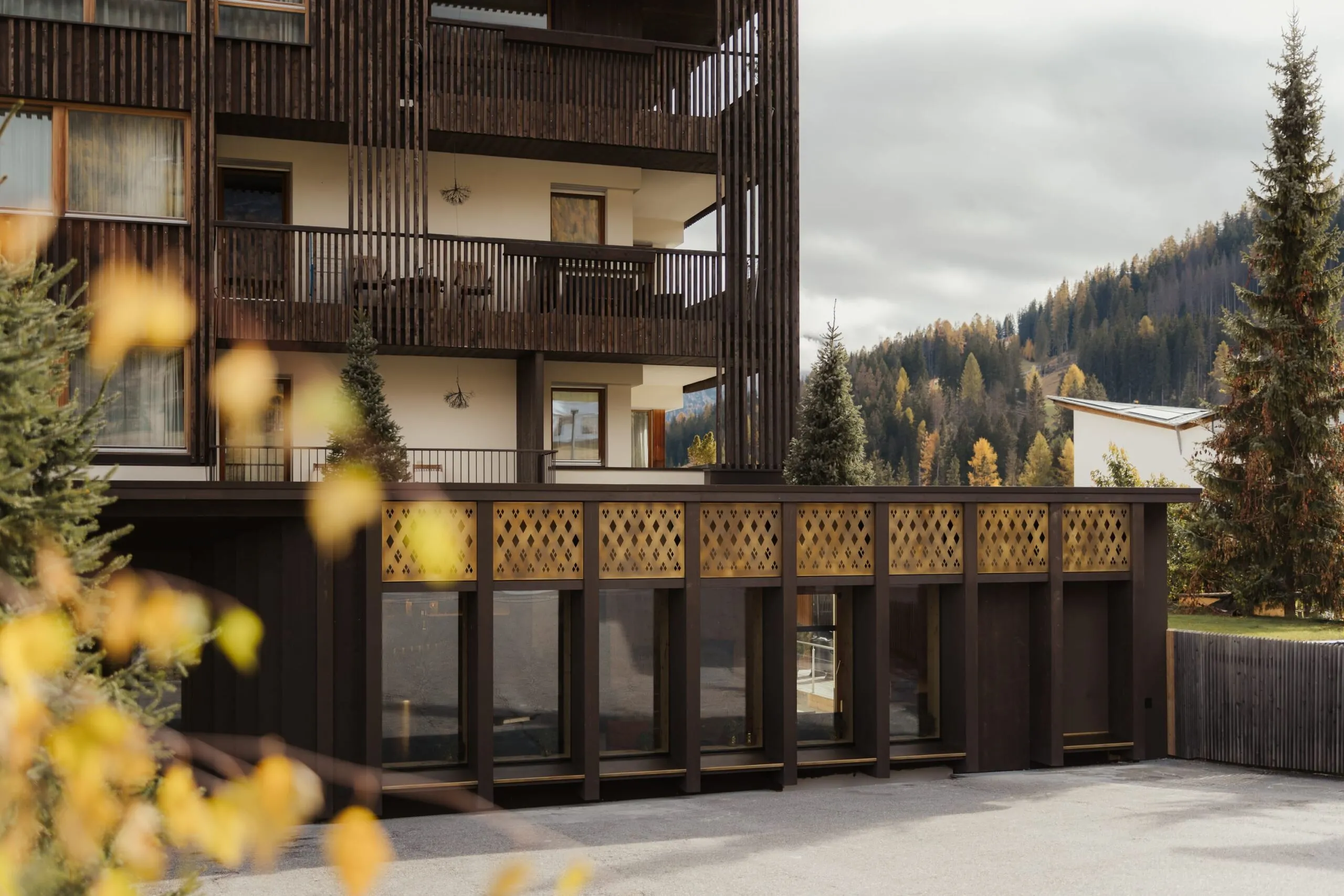 Mehrstöckiges Gebäude mit dunkler Holzfassade und Balkonen; untere Etage mit Gitterverkleidung, davor gelbe Herbstblätter und Wald im Hintergrund.