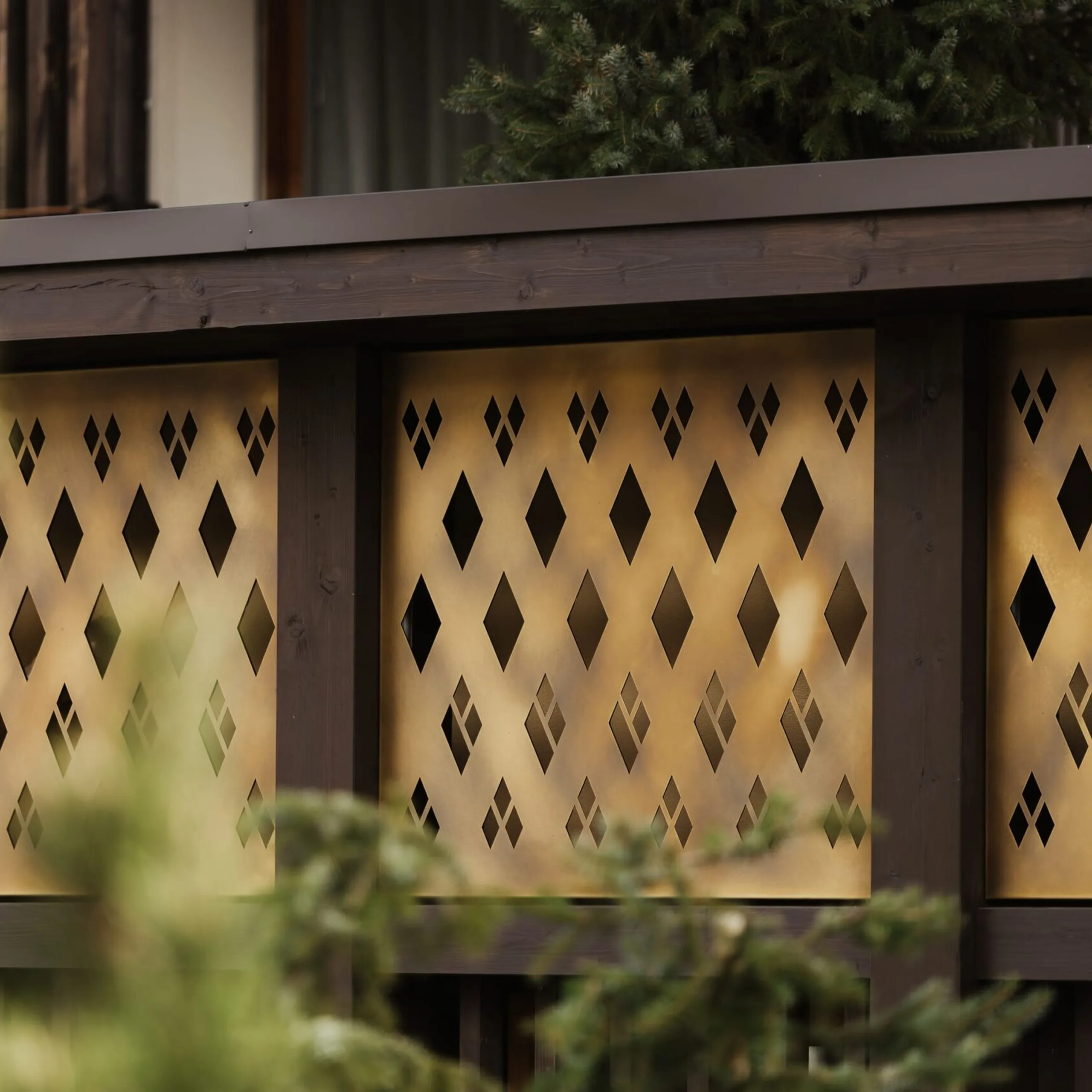 Goldfarbene Metallplatten mit diamantförmigen Ausschnitten, von dunklem Holzrahmen eingefasst; unscharfe grüne Zweige im Vordergrund.