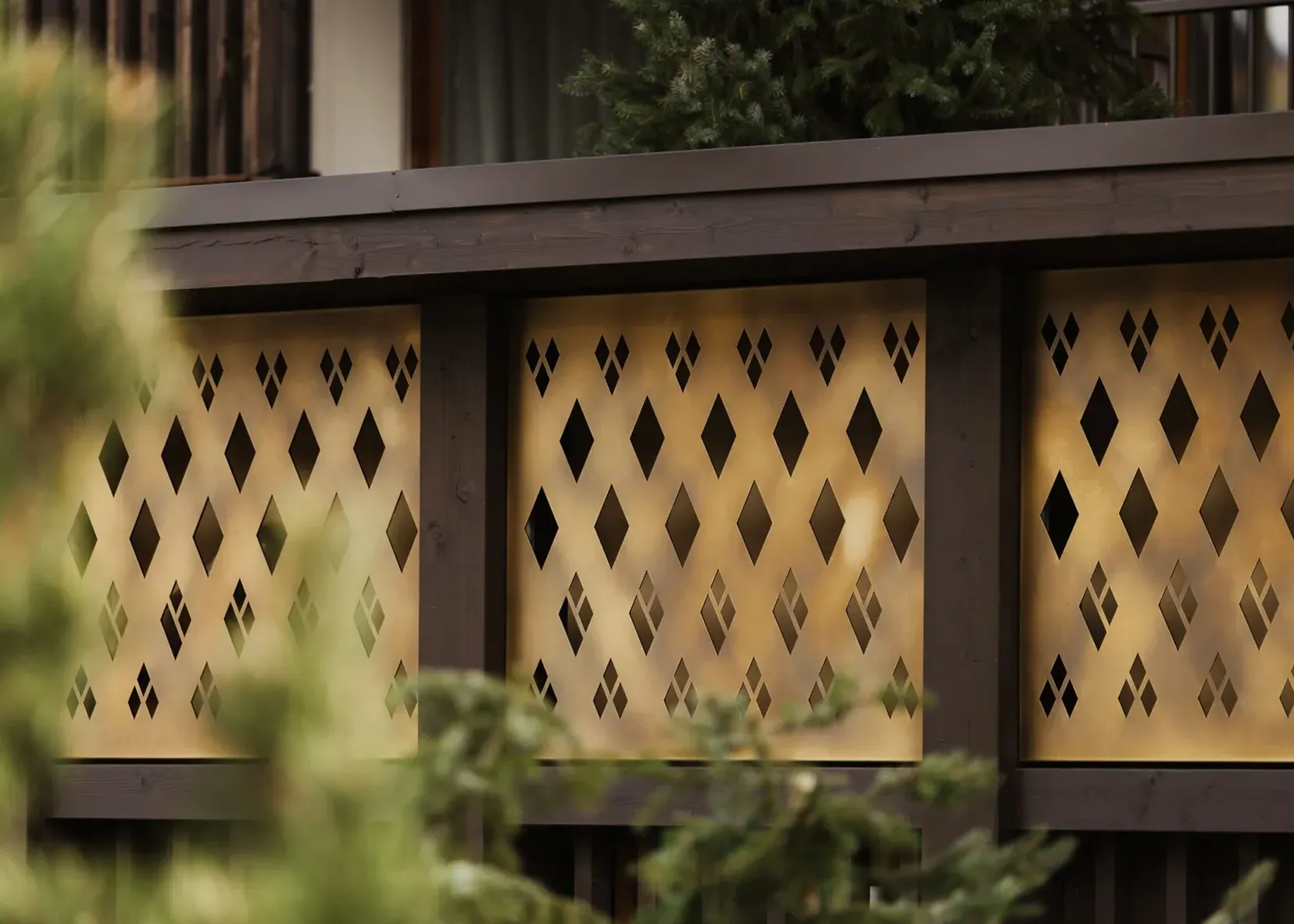 Goldfarbene Metallplatten mit diamantförmigen Ausschnitten, von dunklem Holzrahmen eingefasst; unscharfe grüne Zweige im Vordergrund.