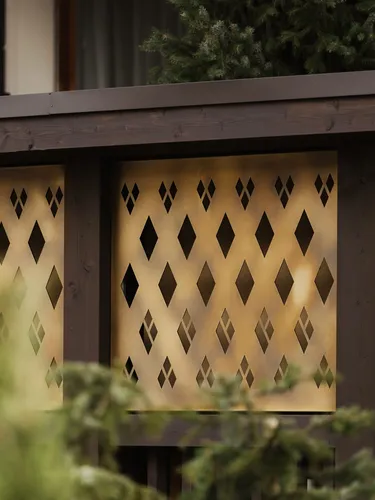 Goldfarbene Metallplatten mit diamantförmigen Ausschnitten, von dunklem Holzrahmen eingefasst; unscharfe grüne Zweige im Vordergrund.