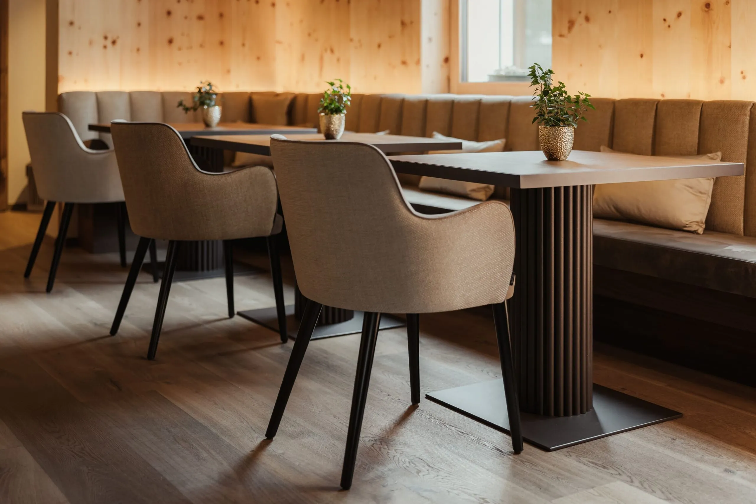Moderner Speisebereich mit beigen, gepolsterten Stühlen, rechteckigen Tischen auf runden Metallstützen, Topfpflanzen auf den Tischen; Holzpaneele und Bankensitz.