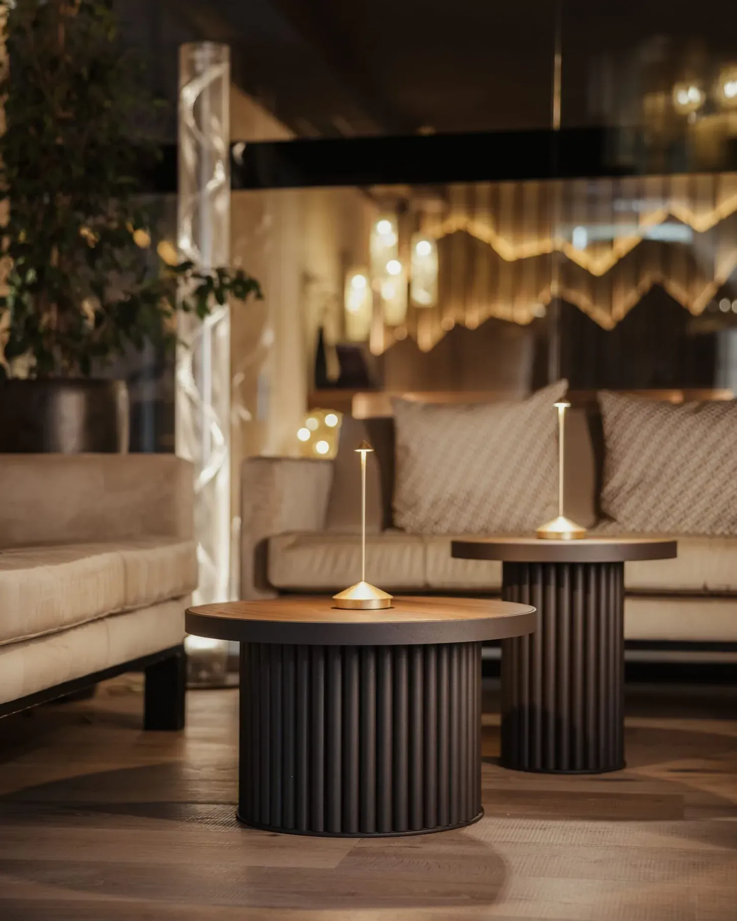 Moderne Lounge mit runden schwarzen Beistelltischen auf geriffelten Basen, Holzplatten, beigen Sofas, Pflanzen und warmem Ambiente-Licht.