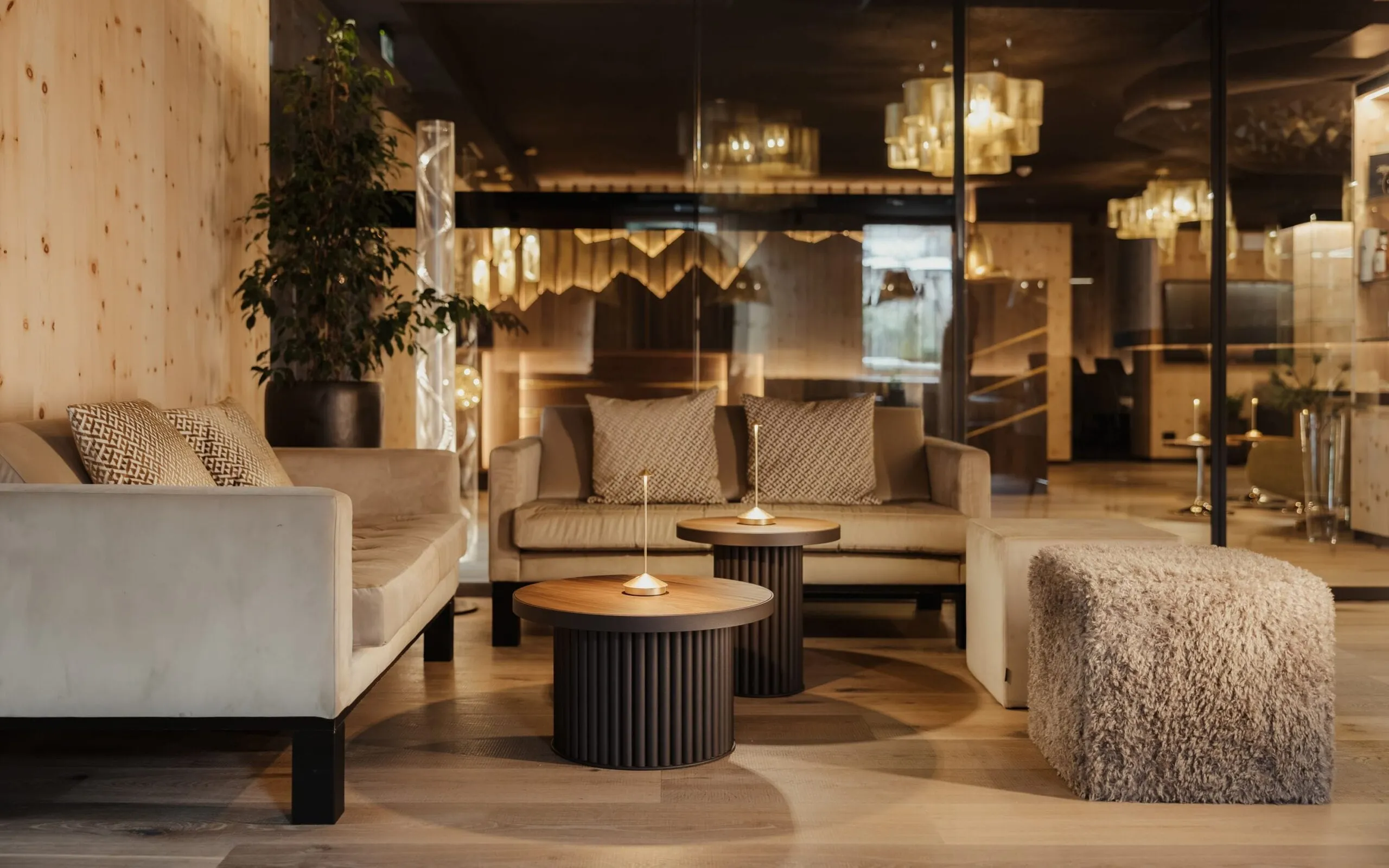 Zona lounge in legno chiaro con divani beige, candele sui tavolini circolari neri, pouf shaggy grigio, pianta e vetrata con luci calde.