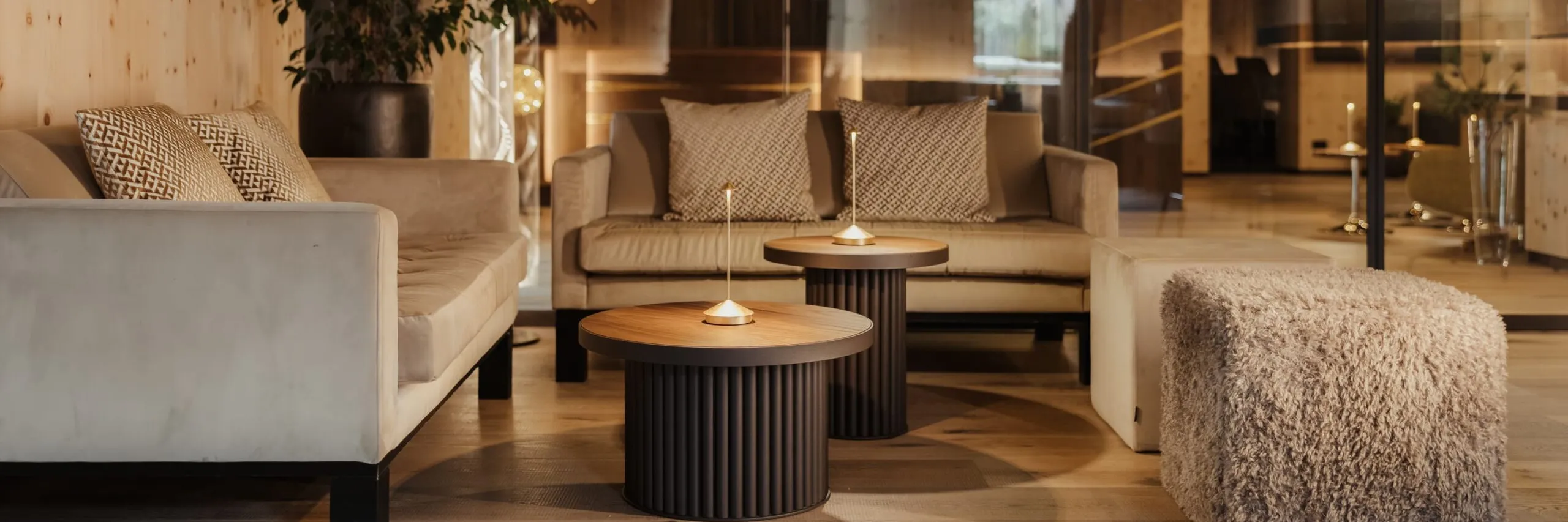 Moderne Lounge eines Metallbaubetriebs in Südtirol: beiges Sofaset, runde Holztische mit Kerzen, ein flauschiger Schemel, Glasfront im Hintergrund.