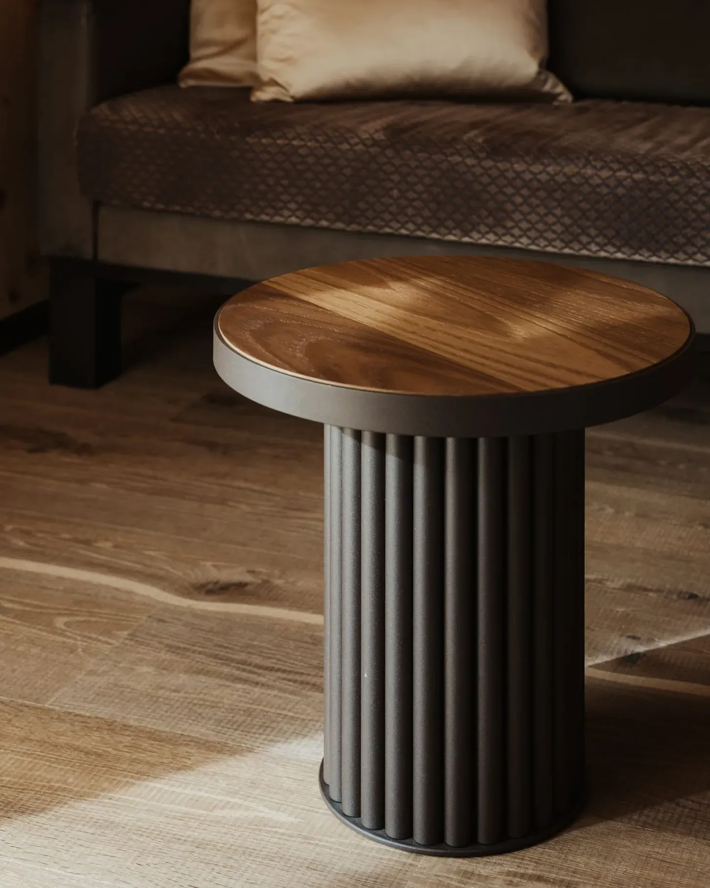 Runder Holztisch mit dunklem, geripptem Metallfuß im Vordergrund; dahinter dunkles Sofa mit hellen Kissen, Holzboden und Holzwand.