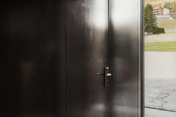Innenaufnahme eines dunklen Metallraums mit einer großen Tür; Licht von außen fällt hinein, Blick nach draußen auf Gras, Gebäude und Berglandschaft.