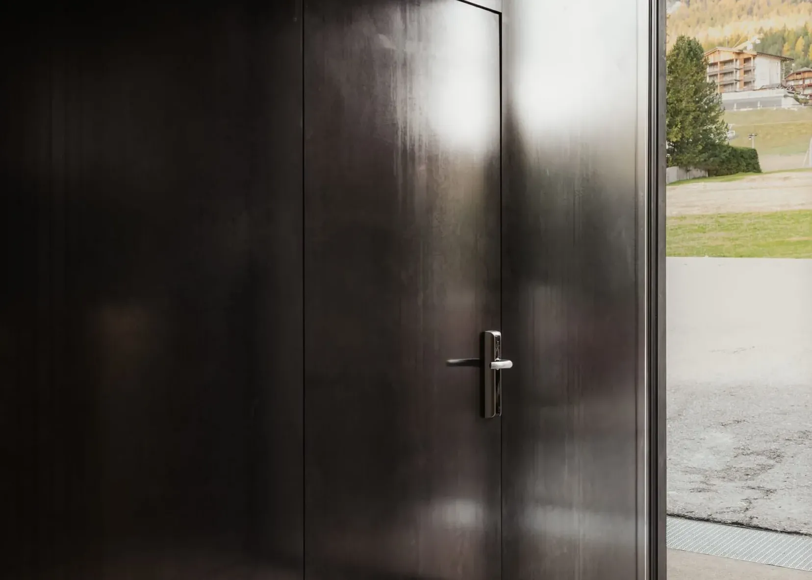 Innenaufnahme eines dunklen Metallraums mit einer großen Tür; Licht von außen fällt hinein, Blick nach draußen auf Gras, Gebäude und Berglandschaft.