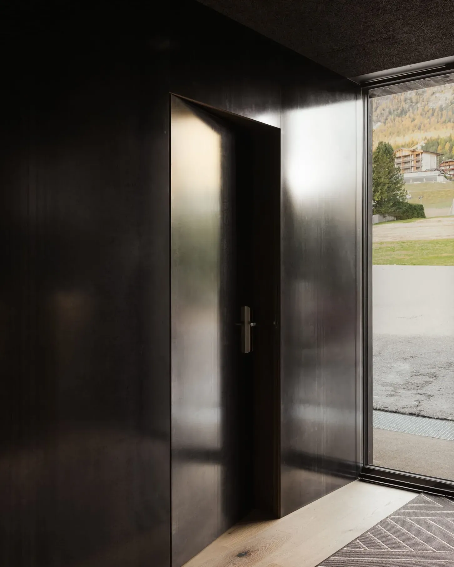 Dunkler Metallraum mit offener Tür zur Außenwelt; Holzboden im Eingangsbereich; Blick auf grüne Südtirol-Landschaft mit Gebäuden am Horizont.