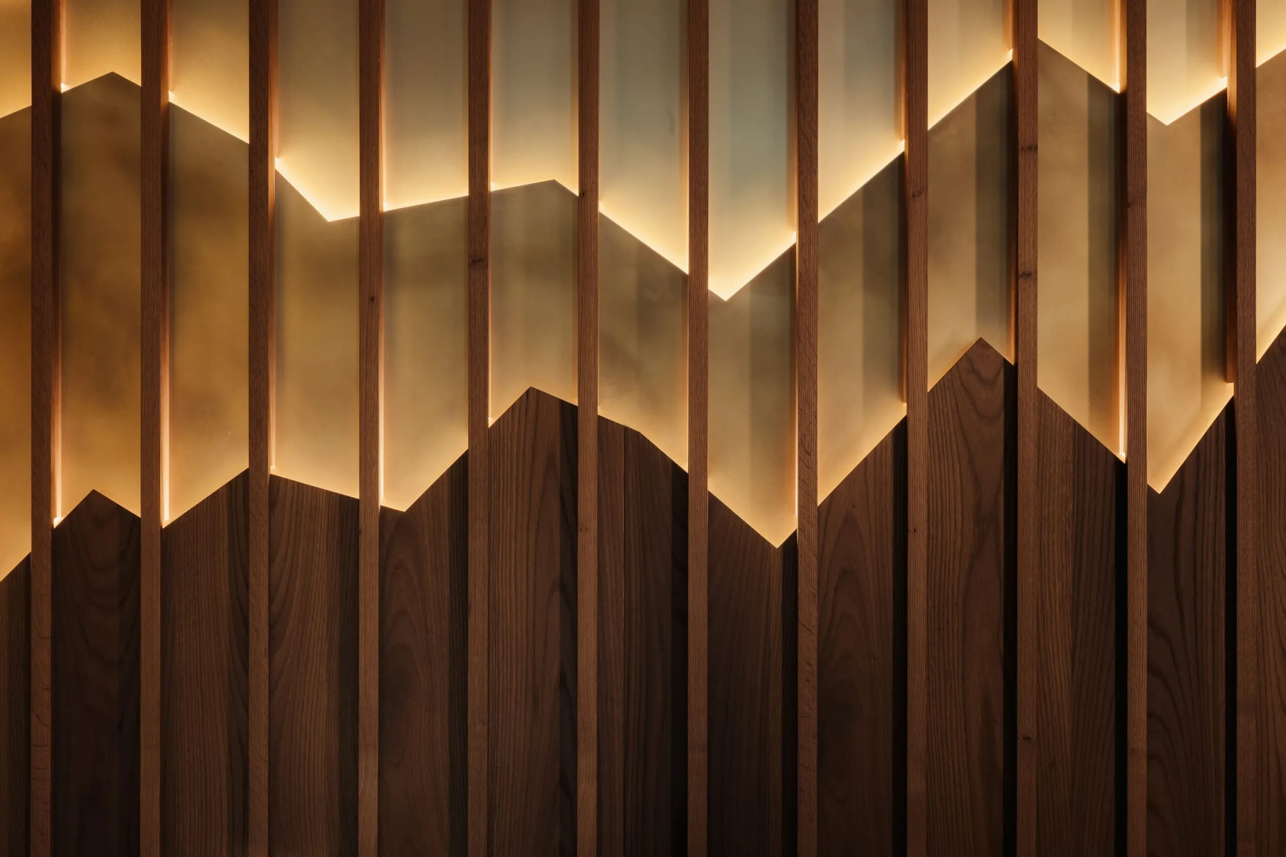 Vertikale Holzbalken vor warm beleuchtetem Hintergrund, der chevronartige Gebirgskanten bildet; unten dunkle Holzpaneele.