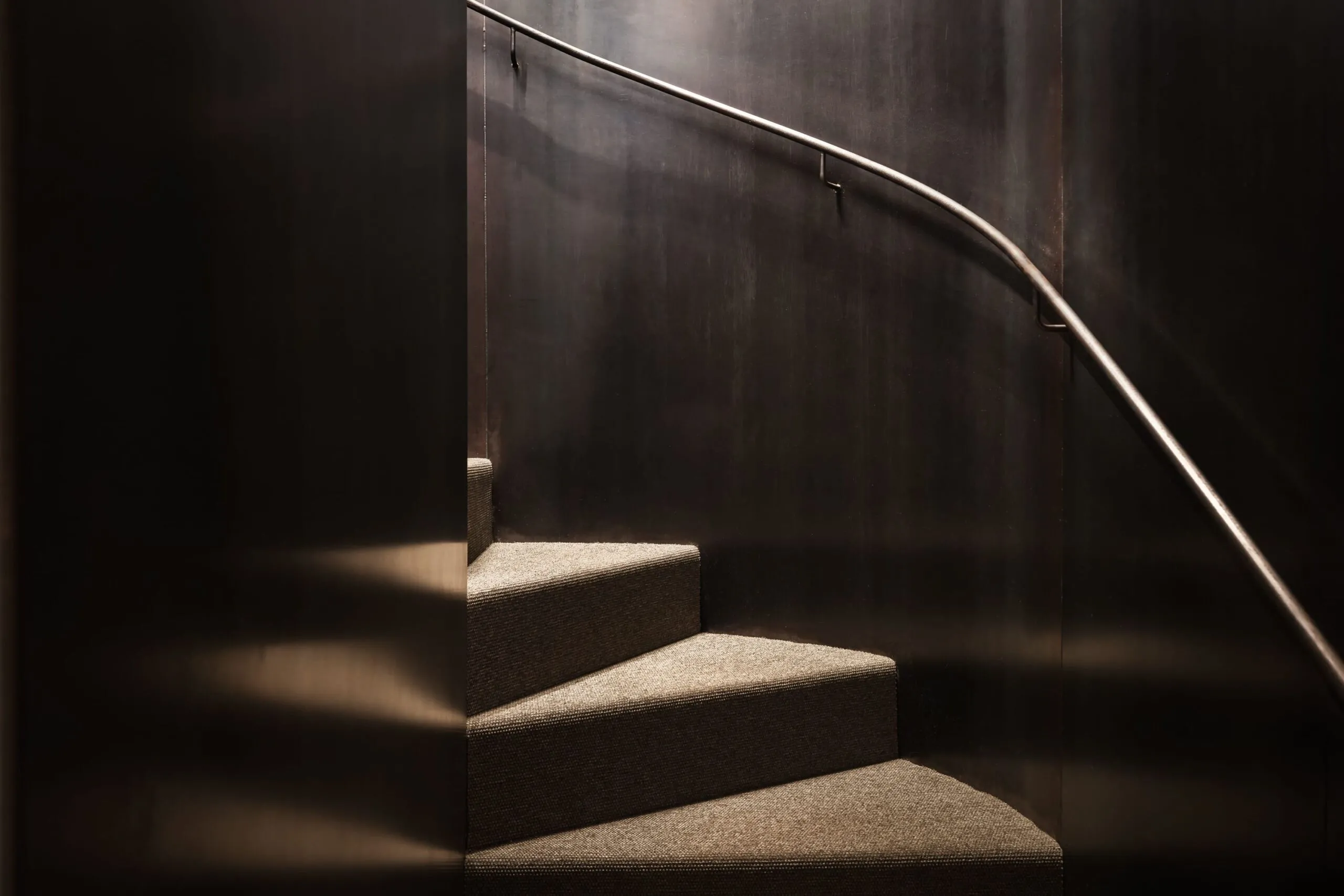 Scalinata interna in stile industriale: gradini rivestiti di moquette grigia, pareti metalliche scure e corrimano in metallo curvo lungo il lato.