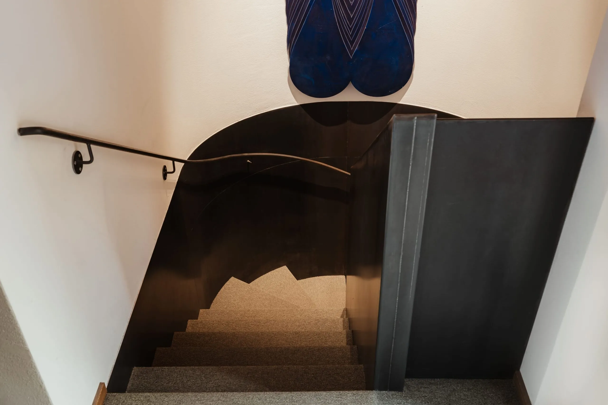 Scalinata interna con corrimano a sinistra, parete metallica nera riflettente curva a destra, gradini in moquette; figura umana vestita di blu in alto.