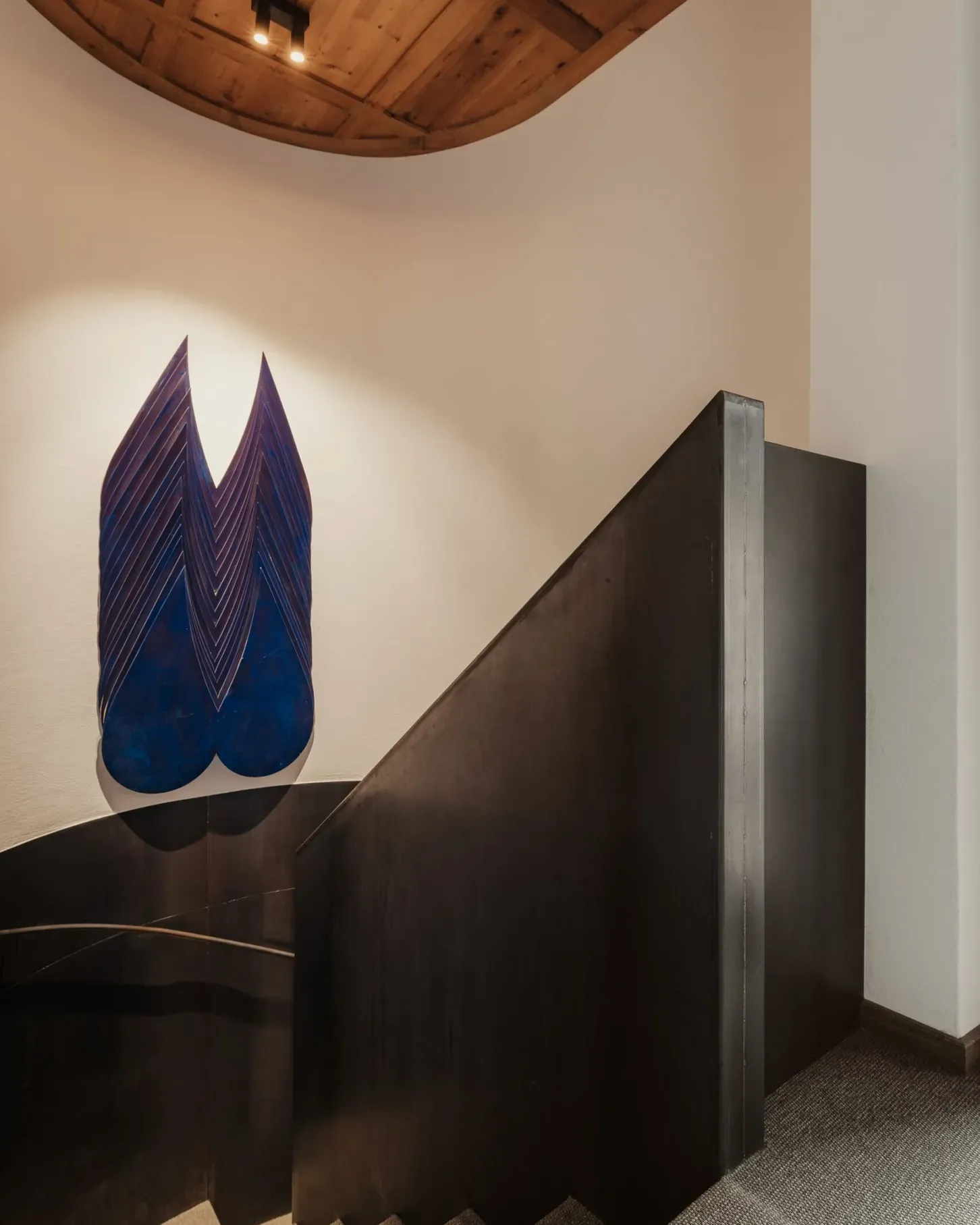 Interno con scala a chiocciola in legno scuro e parete bianca. Scultura blu a ali sovrapposte; soffitto circolare in legno e illuminazione a spot.