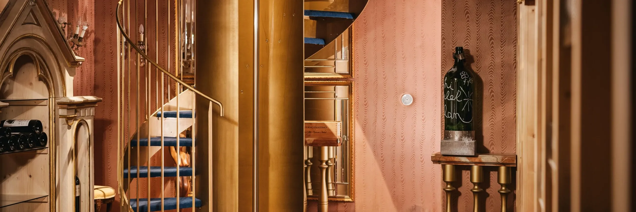 Interno di un’azienda metalmeccanica in Südtirol: scala a chiocciola dorata al centro, pareti rosa, pavimento in legno; bottiglia su tavolo a destra.