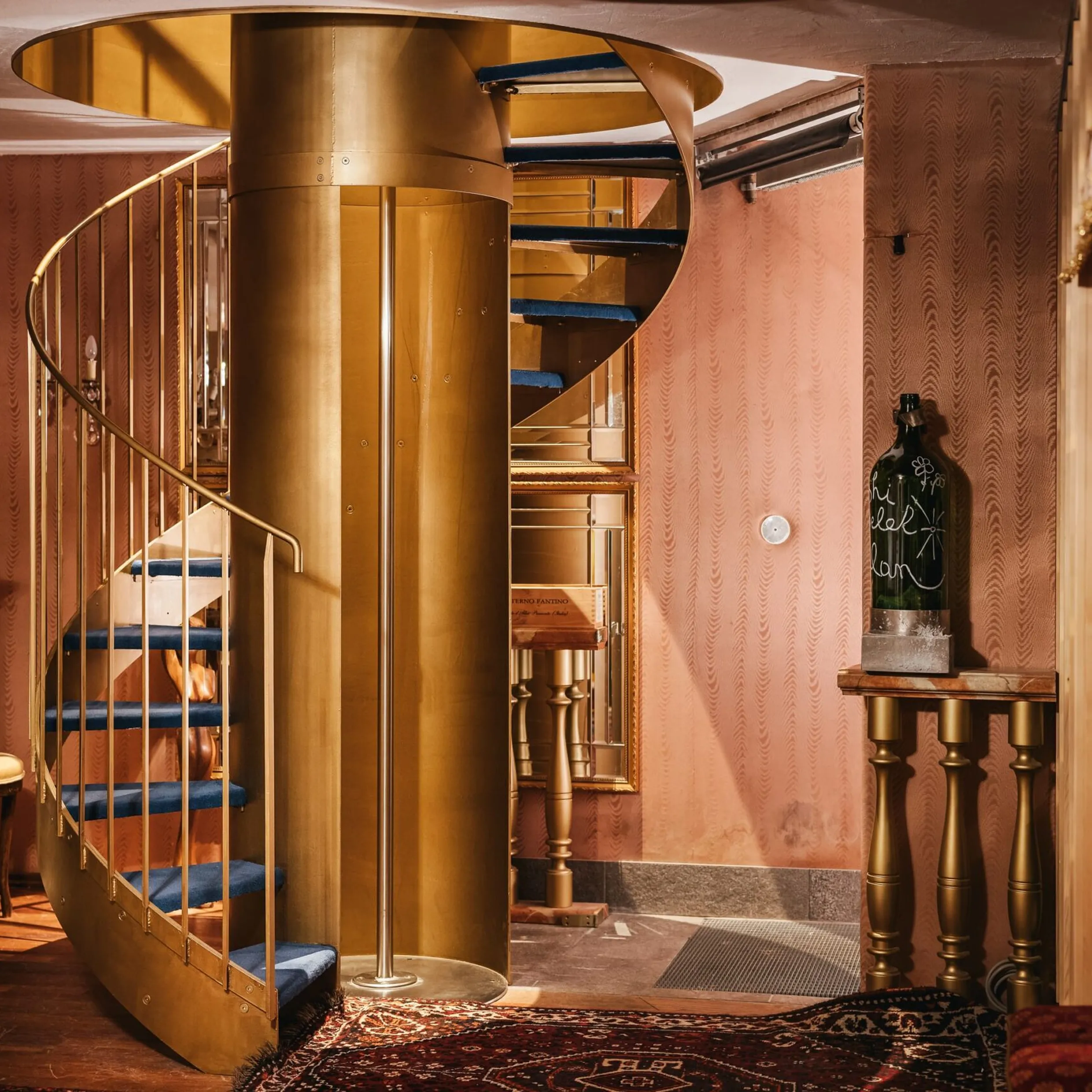 Interno di un’azienda metalmeccanica in Südtirol: scala a chiocciola dorata al centro, pareti rosa, pavimento in legno; bottiglia su tavolo a destra.