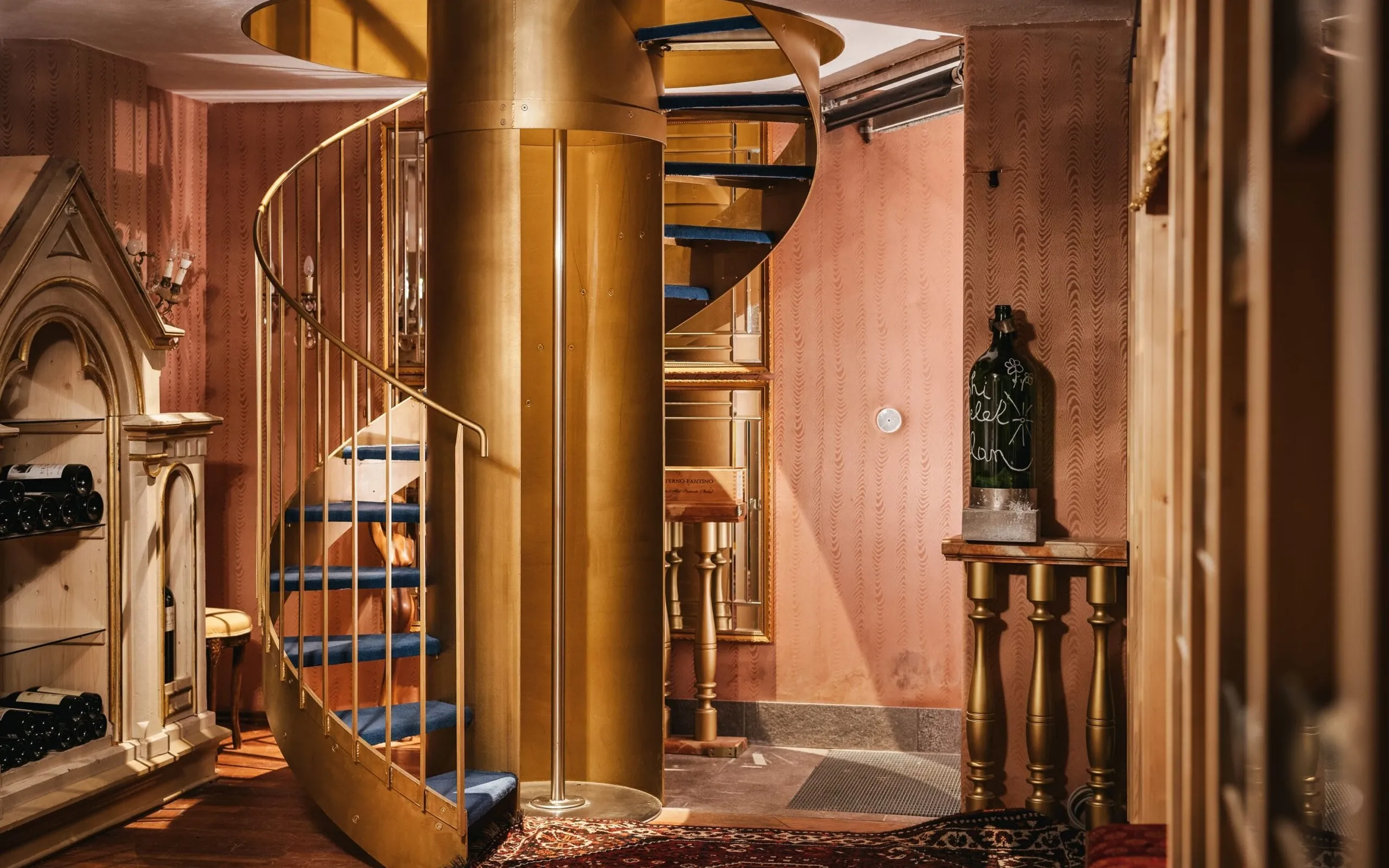 Interno di un’azienda metalmeccanica in Südtirol: scala a chiocciola dorata al centro, pareti rosa, pavimento in legno; bottiglia su tavolo a destra.