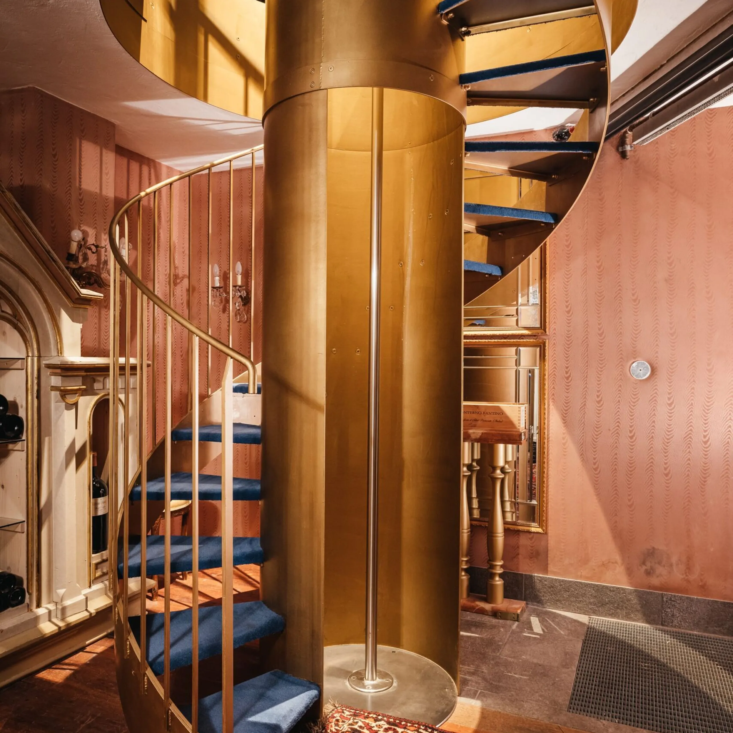 Scaletta a chiocciola in ottone dorato con gradini blu, corrimano dorato; interno elegante con pareti rosa, tappeto persiano e bottiglie sul ripiano superiore.