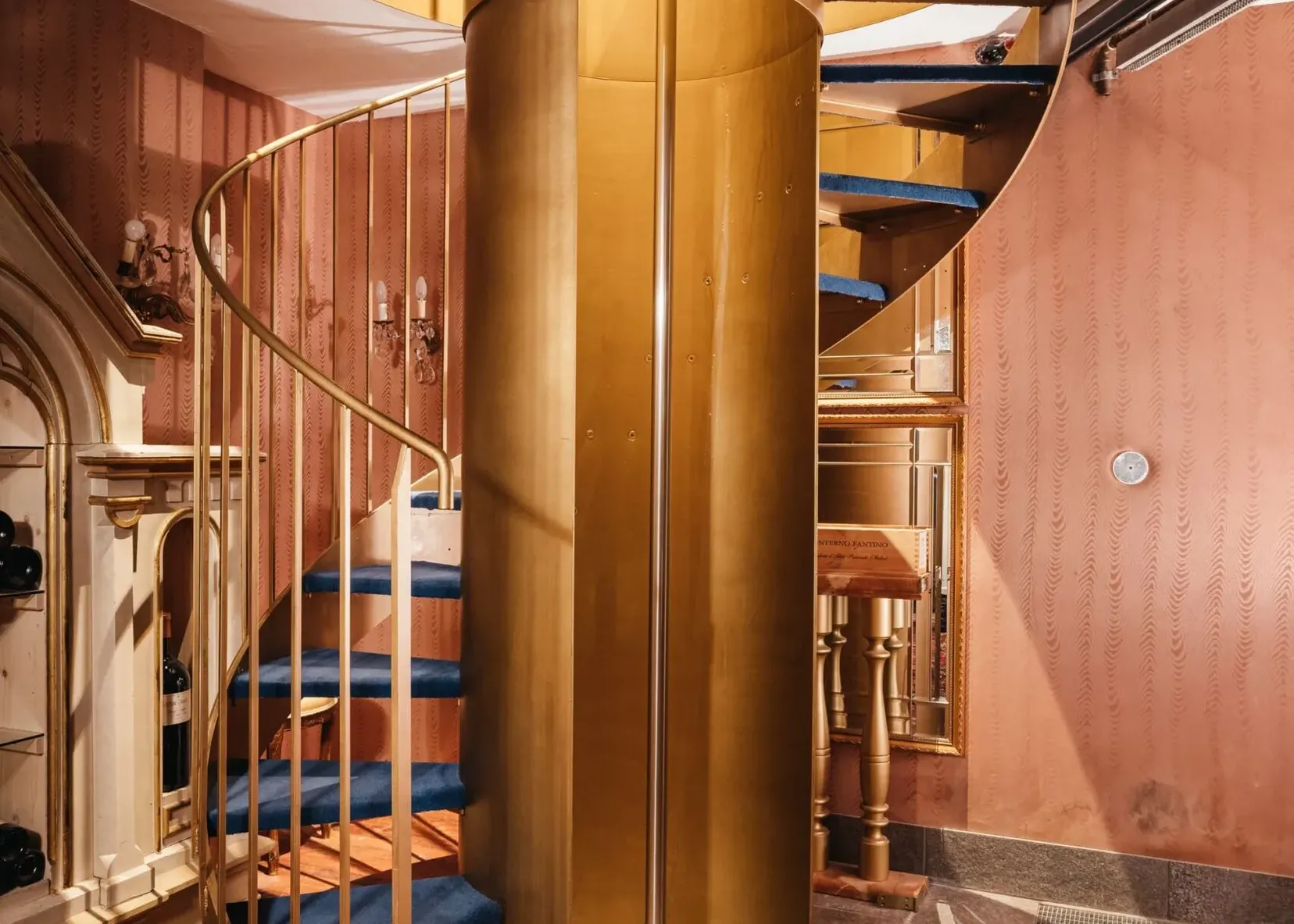 Scaletta a chiocciola in ottone dorato con gradini blu, corrimano dorato; interno elegante con pareti rosa, tappeto persiano e bottiglie sul ripiano superiore.