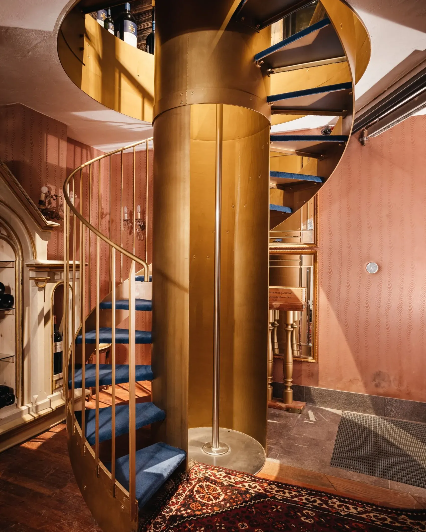 Scaletta a chiocciola in ottone dorato con gradini blu, corrimano dorato; interno elegante con pareti rosa, tappeto persiano e bottiglie sul ripiano superiore.