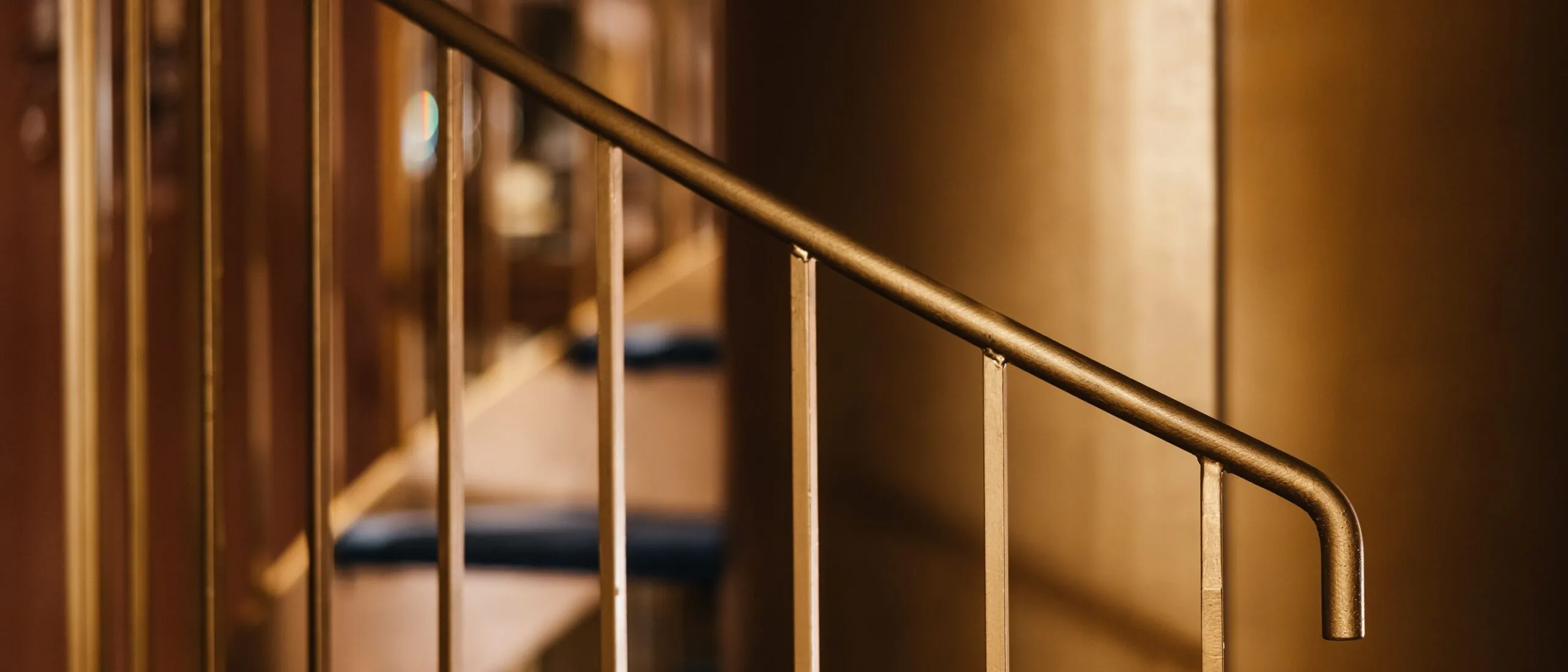 Primo piano su una ringhiera in metallo con corrimano curvo e barre verticali, illuminazione calda che permea lo spazio, sfondo sfumato.