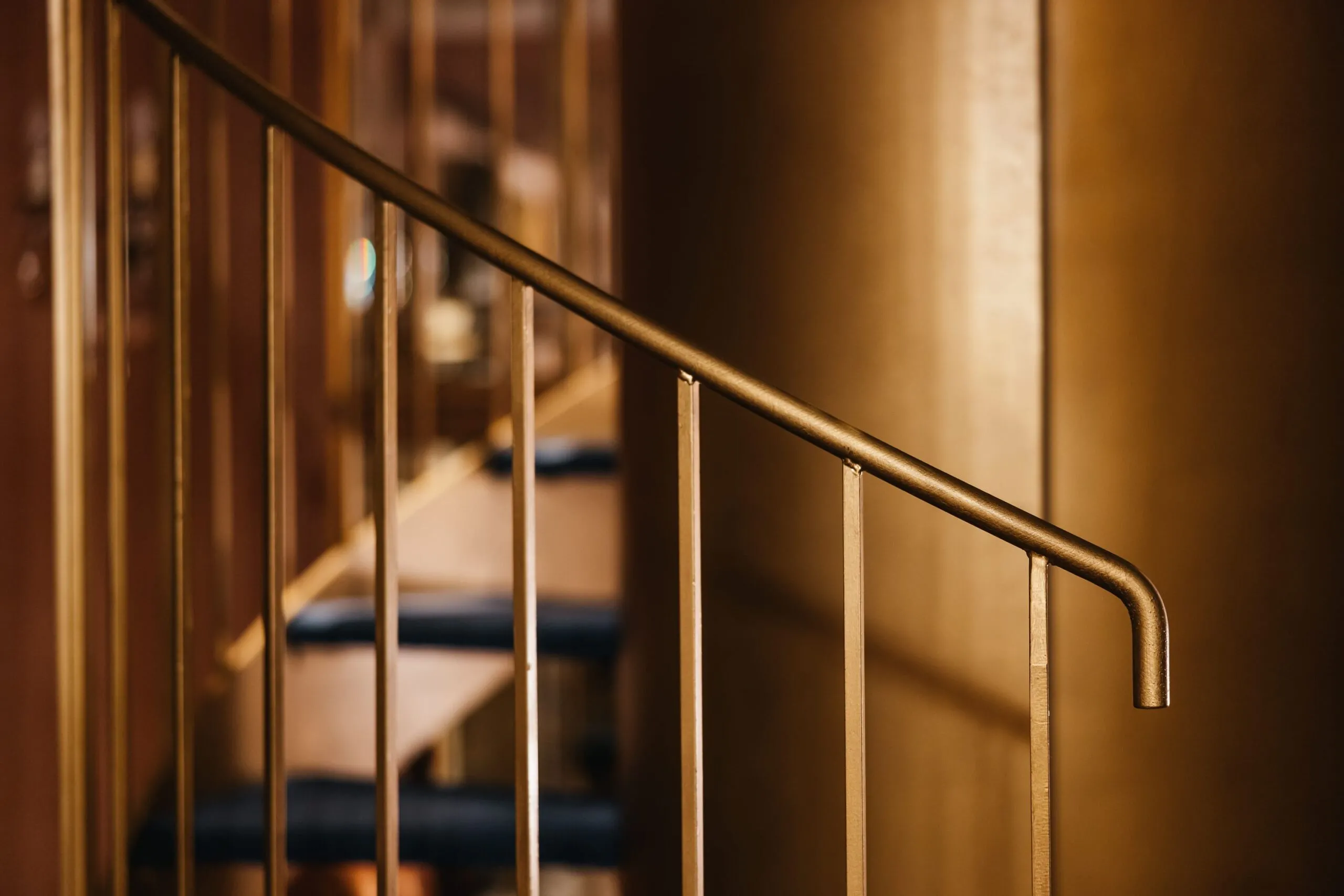Primo piano su una ringhiera in metallo con corrimano curvo e barre verticali, illuminazione calda che permea lo spazio, sfondo sfumato.