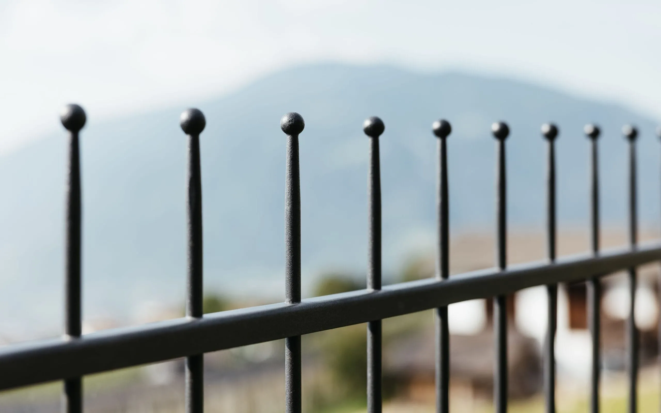 Primo piano di una recinzione in metallo con barre verticali a punte tonde; sfondo sfocato di montagne e case nell'Alto Adige.